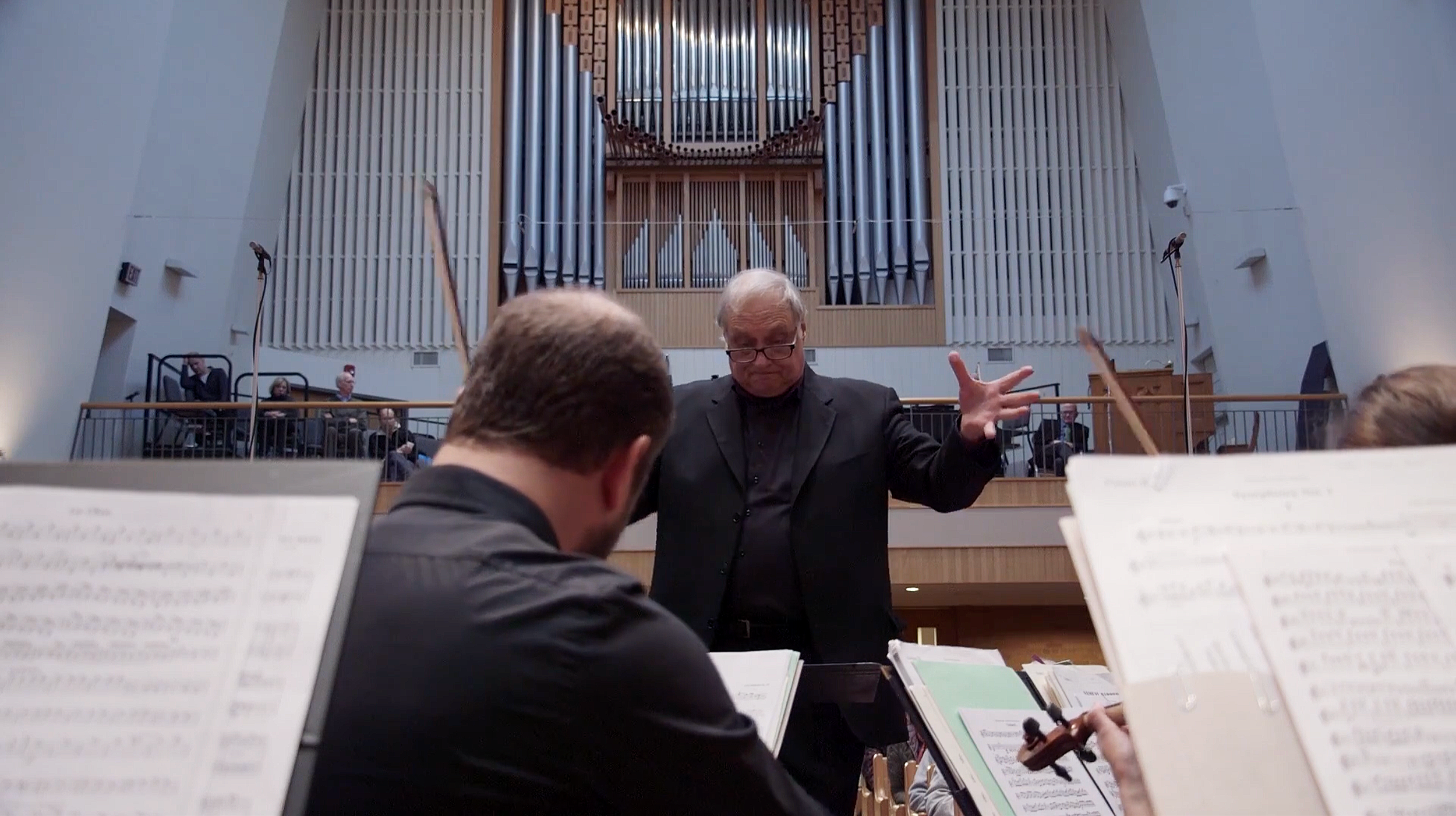 A conductor leading an orchestra during a performance in a church or concert hall with a pipe organ in the background.