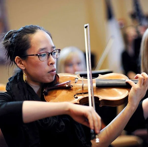 A woman with glasses and earrings playing the violin in an orchestra rehearsal.