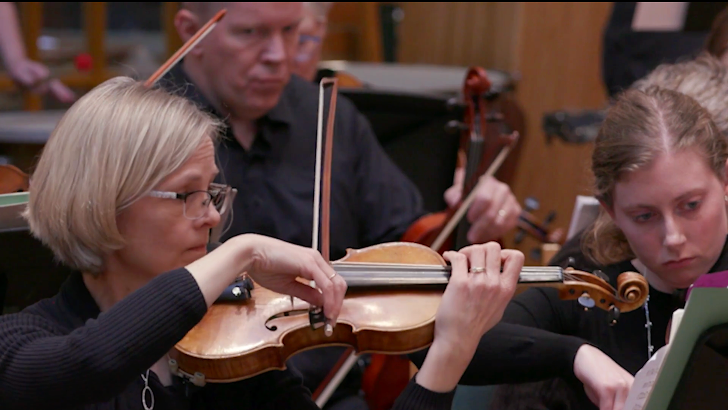 Group of musicians playing string instruments in an orchestra, including a woman with glasses and a woman with light brown hair, focused on sheet music.