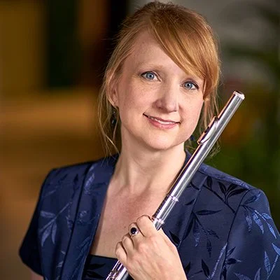 Red-haired woman smiling, wearing a dark blue blazer, holding a silver flute, with a blurred indoor background.