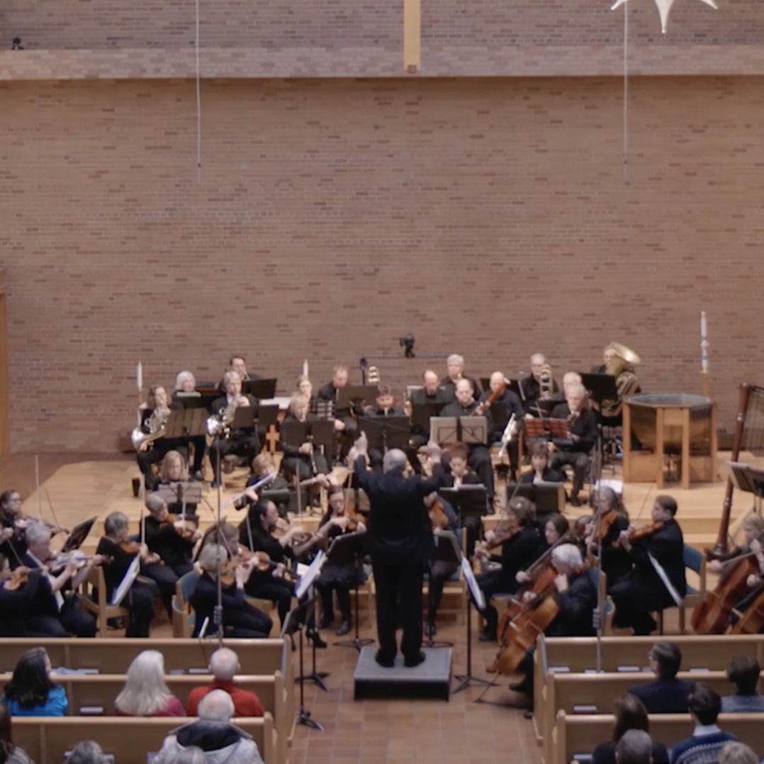 Orchestra performing on stage in a concert hall, led by a conductor, with audience seated in front.