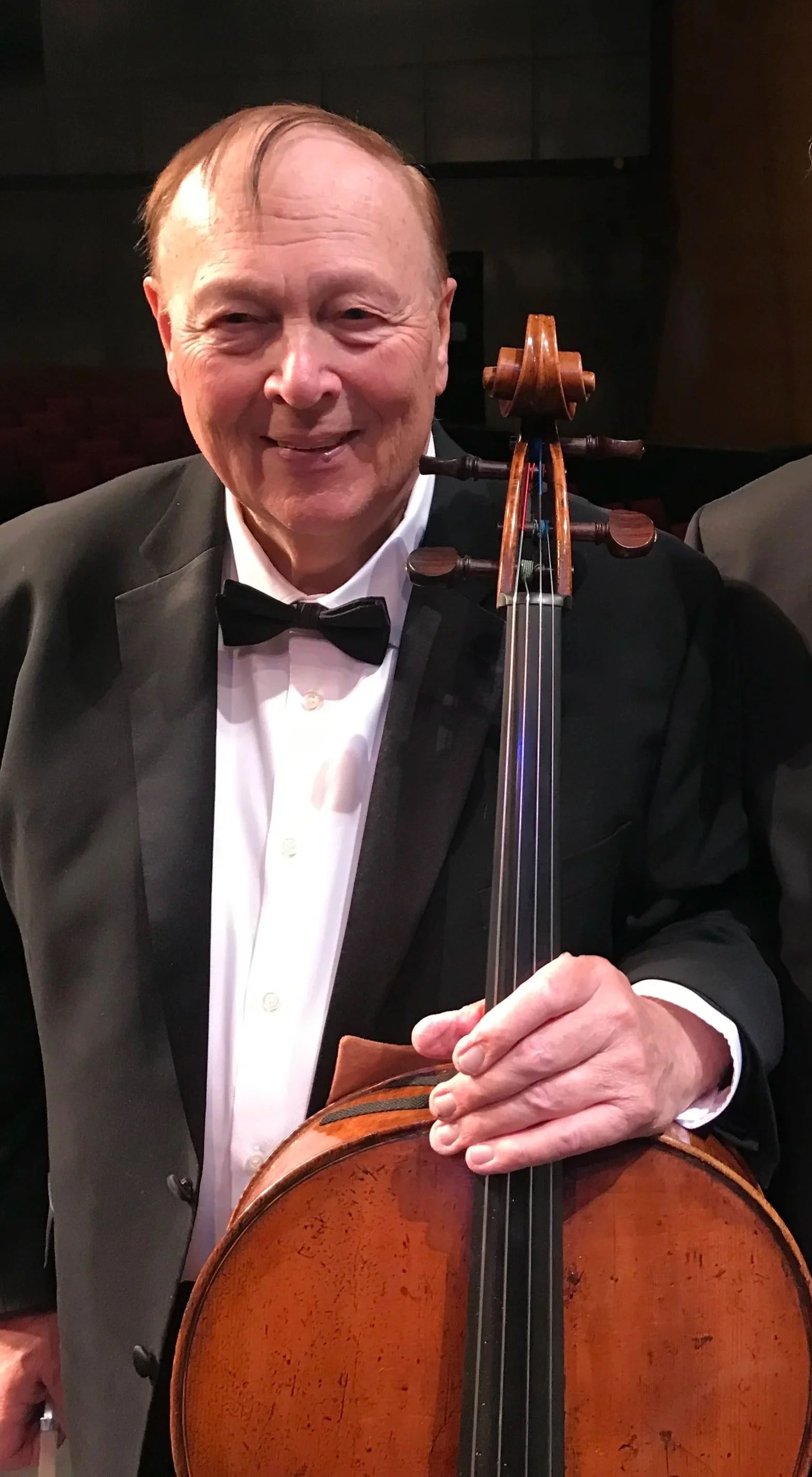 An elderly man in a tuxedo and bowtie holding a violin, smiling, in a dimly lit indoor setting.