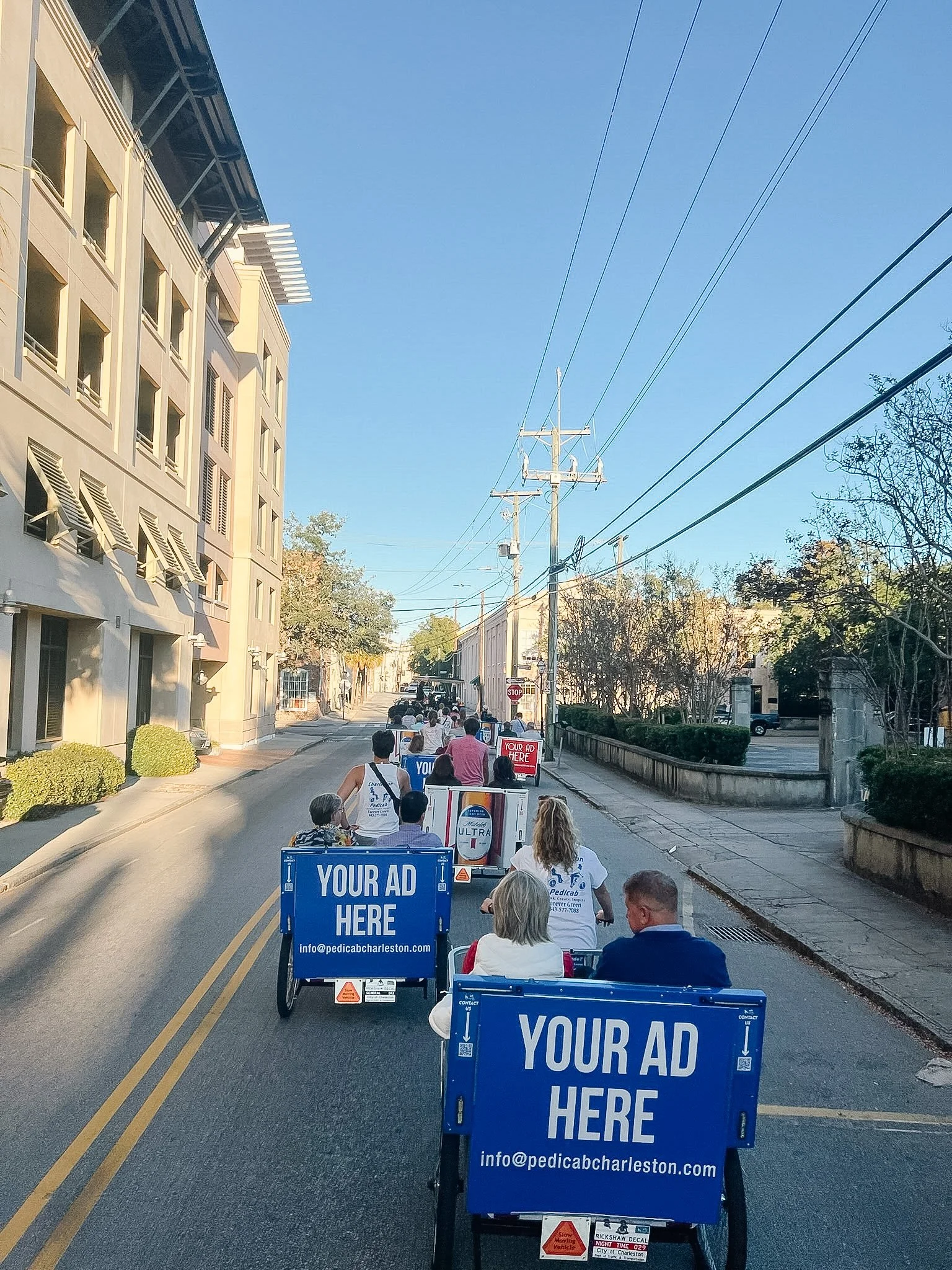 A parade with people riding in bike taxis down a city street in Charleston, SC, advertising space on the carts for ads and contact info, with buildings and power lines lining the street.