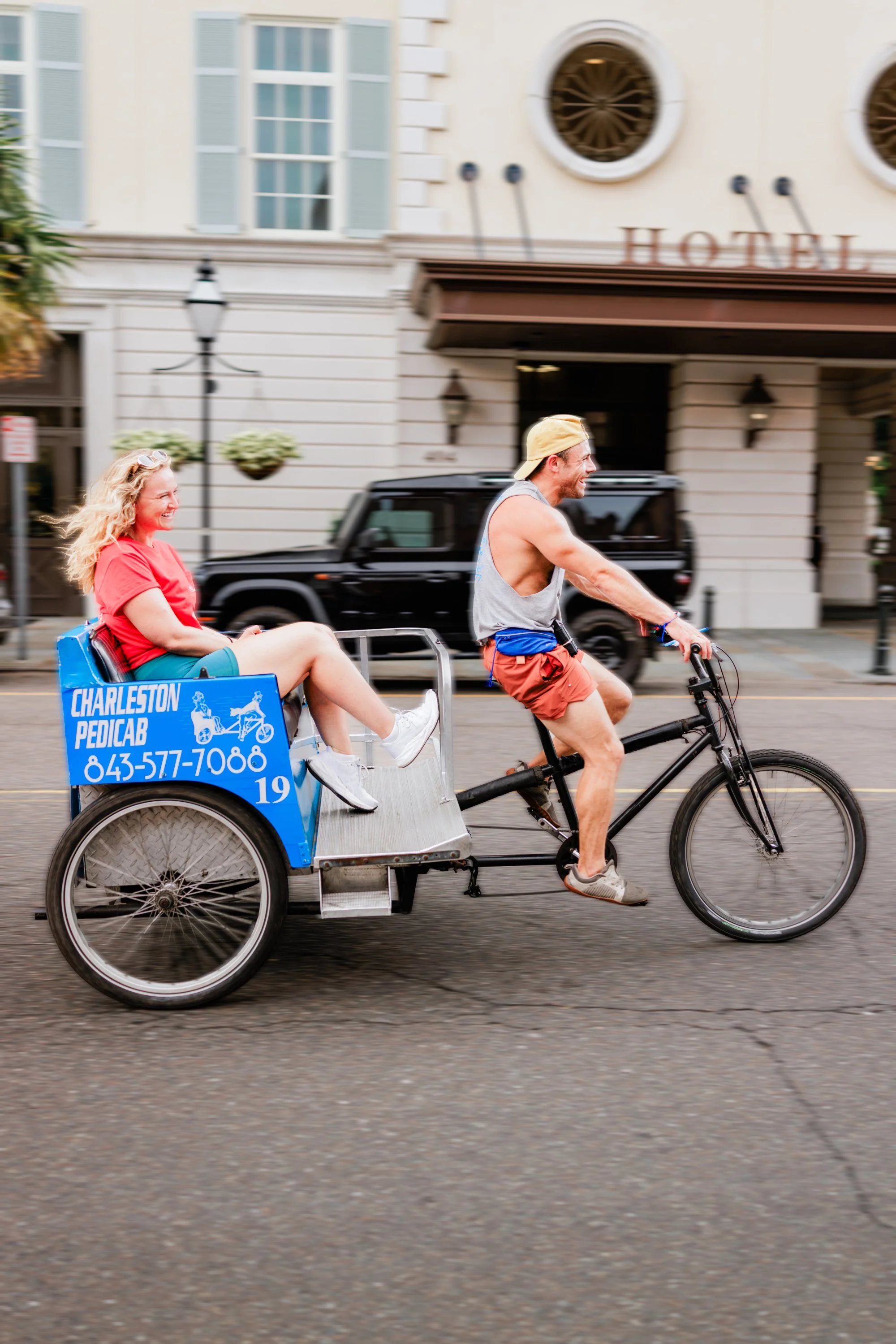 A man driving a bike taxi on a city street in Charleston, SC. The woman is smiling and wearing a red shirt, while the man is wearing a tank top, shorts, and a cap.
