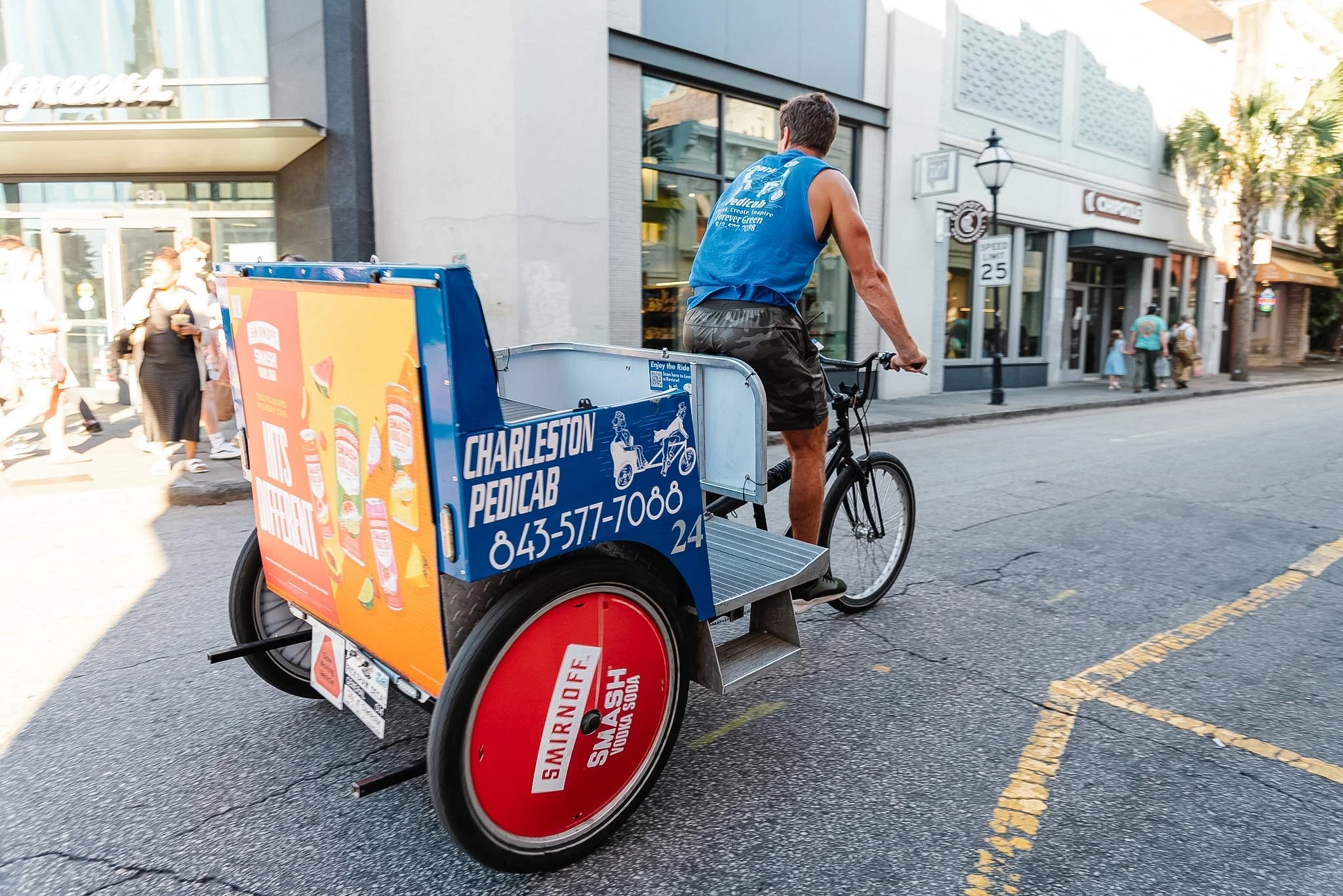 A man riding a bicycle pedicab on a city street, with a brightly colored advertisement for a pedicab service on the side, alongside a sidewalk with pedestrians and storefronts in the background.