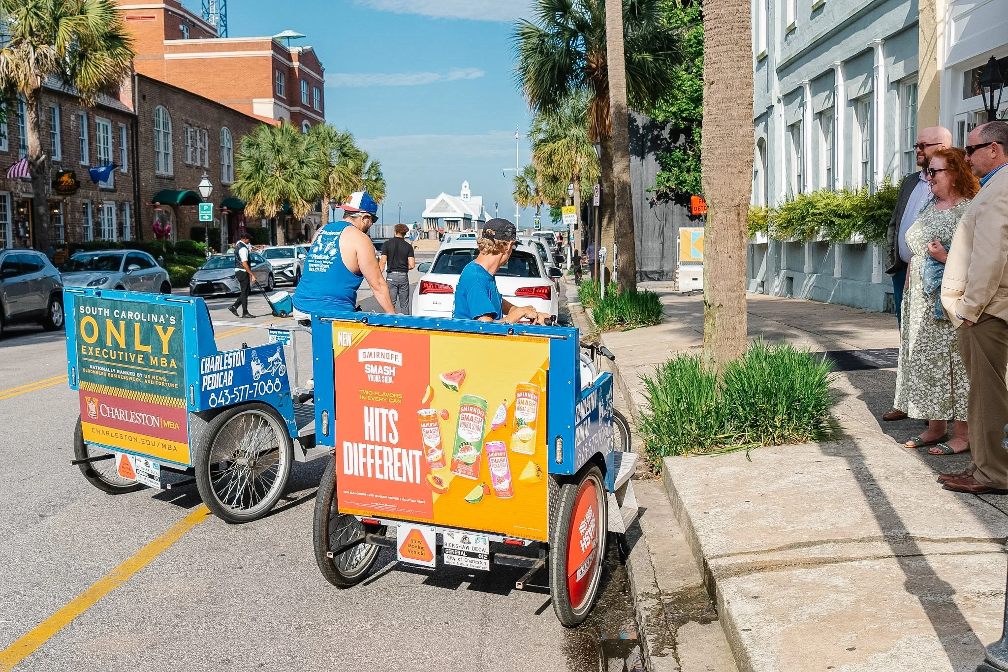 A street scene with a bicycle taxi advertising drinks, with people standing and walking, palm trees, older buildings, and a building with white architecture in the background in Downtown Charleston, SC