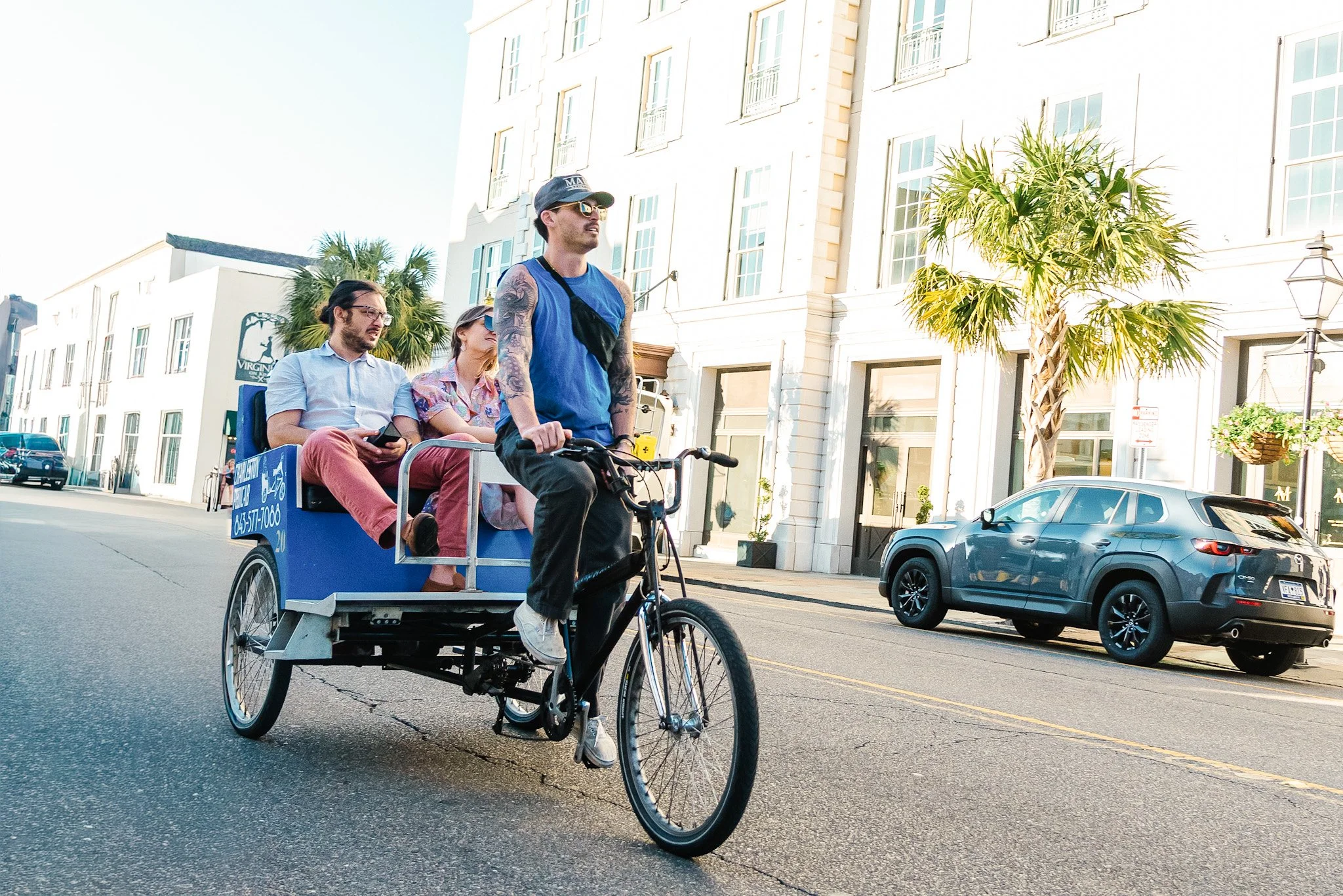 A man rides a bike while pulling a bike taxi with three passengers on a city street in Charleston, SC, with a white building and palm trees in the background.