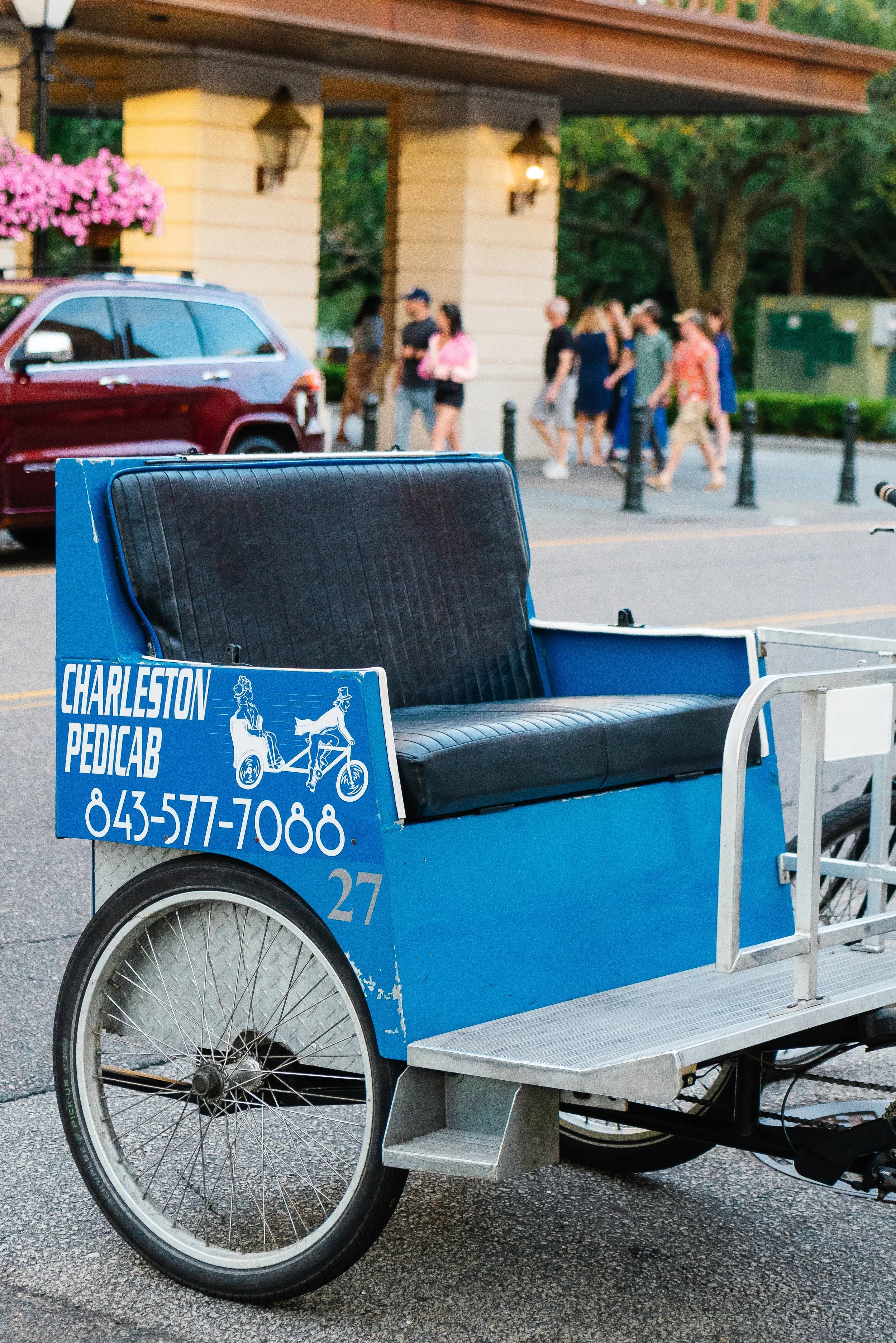A blue pedicab with a black cushioned seat parked on the street. It has advertisements for Charleston Pedicab with a phone number. The background shows a group of people walking outside a building with yellow columns and hanging lanterns.