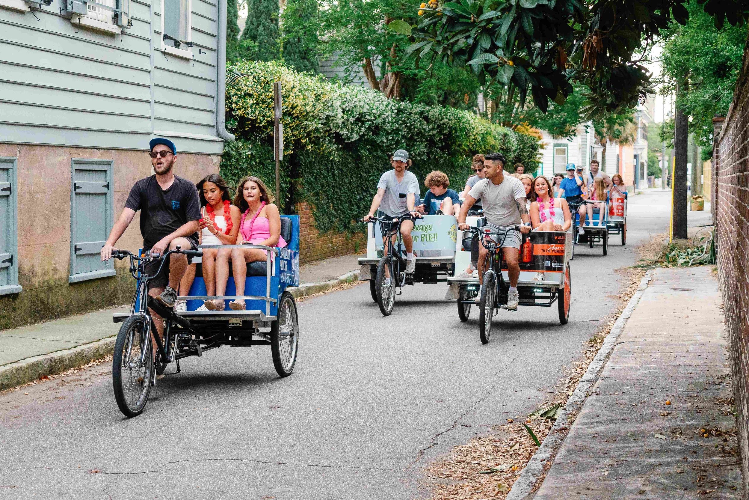 A group of people riding in bike taxis on a narrow residential street in Charleston, SC surrounded by green trees and houses.