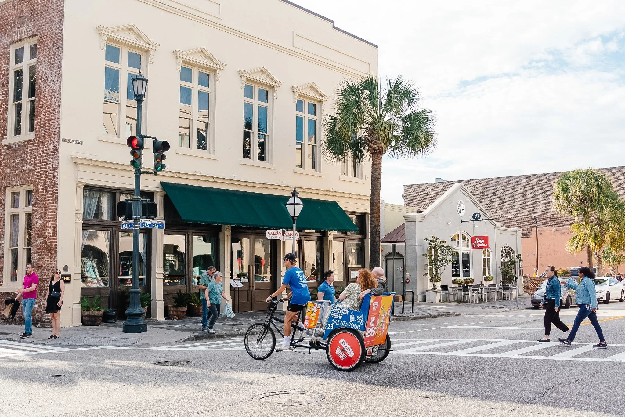 Street scene with people walking and riding in a bike taxi in front of shops, including a restaurant with a green awning. There are palm trees and a traffic light showing red and green signals.