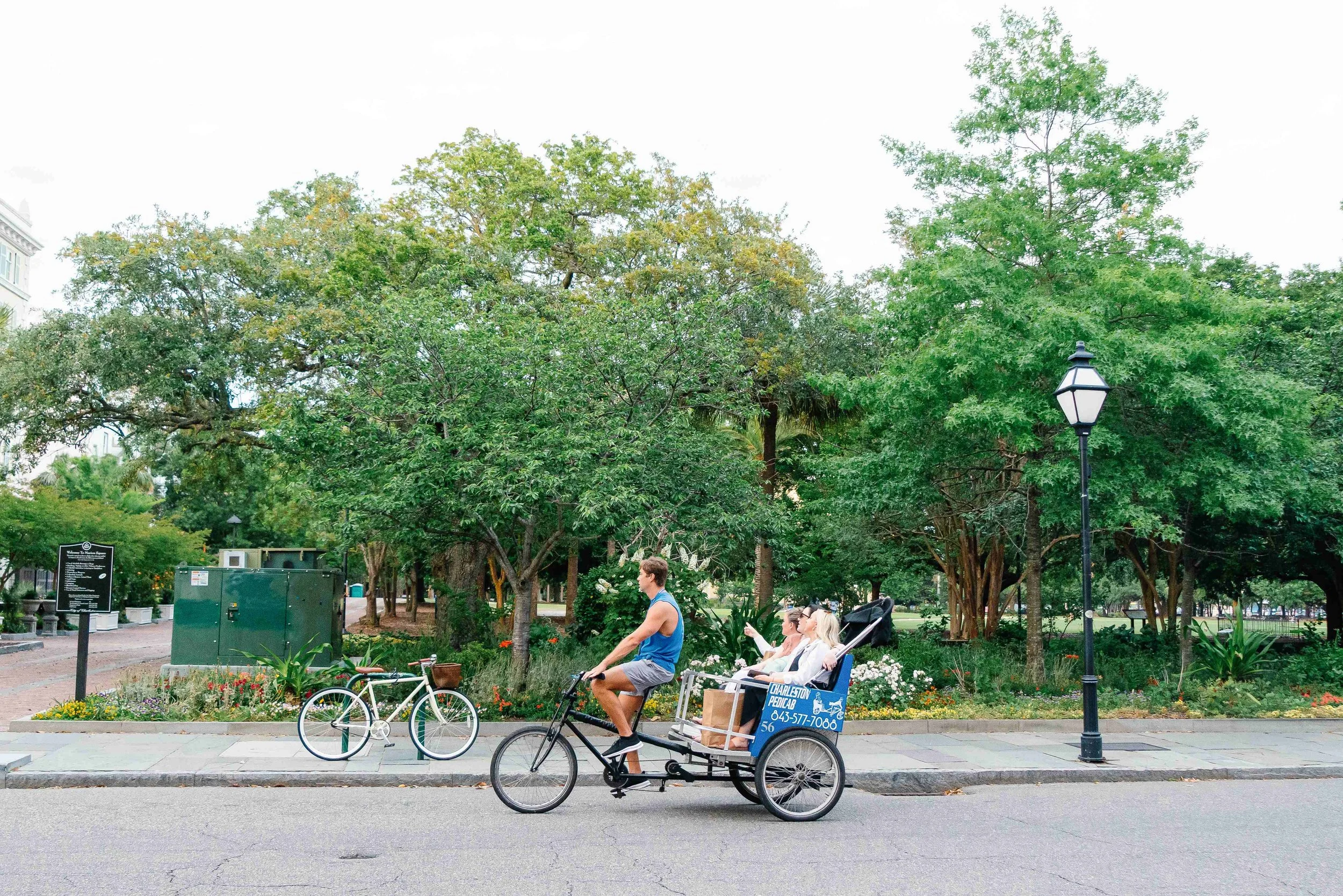 A man riding a bicycle taxi with two women sitting inside in a park with trees, lampposts, and flower beds.