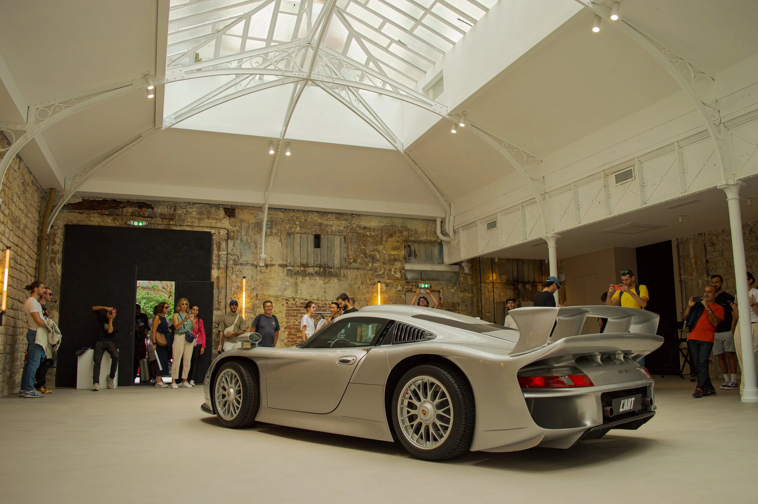 A crowd of people gathered inside a modern building with a high glass ceiling and exposed brick walls, observing a silver sports car, specifically a Porsche 911 GT1, on display.