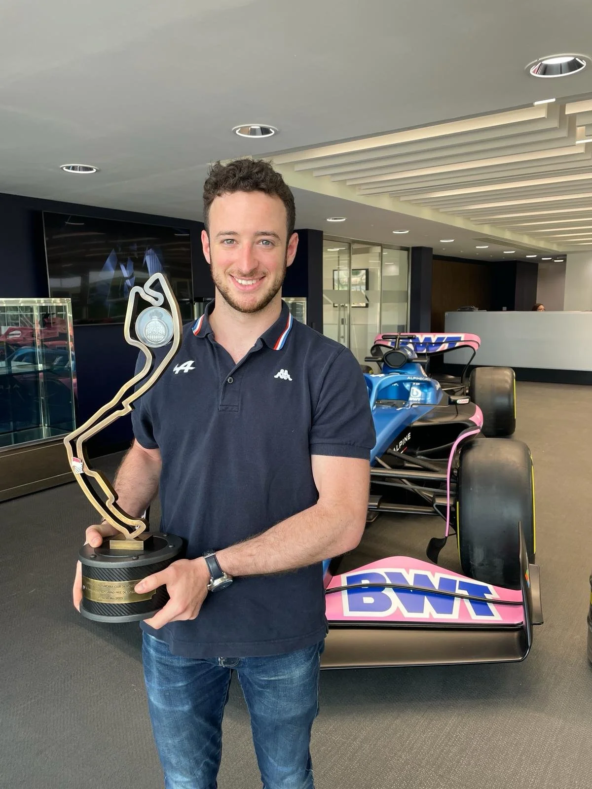 A young man with short curly brown hair and a beard, smiling and holding a trophy in front of a Formula race car, with a modern indoor setting in the background.