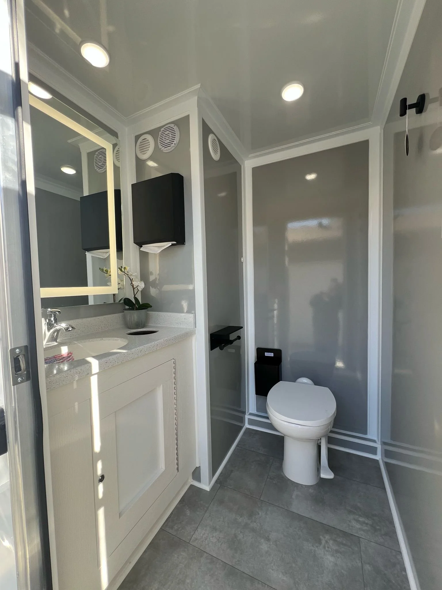 Small bathroom with a white vanity, a mirror, and a toilet. The walls are painted gray, and there are ceiling lights and a wall-mounted black tissue holder. The floor is tiled with gray tiles.
