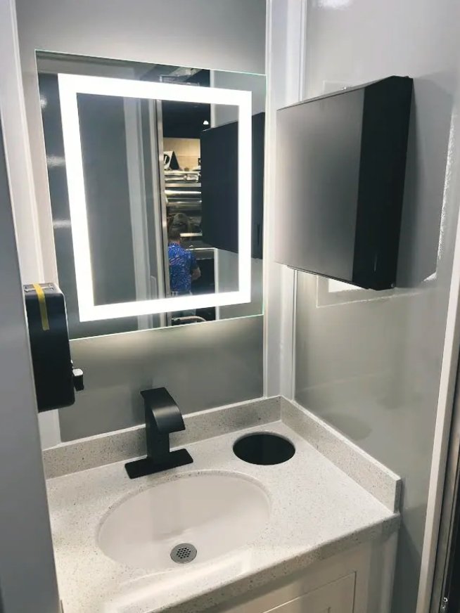 Bathroom sink with a black faucet, a hole for a hand dryer or soap dispenser, a mirror with a built-in light, a wall-mounted paper towel dispenser, and a wall-mounted television.