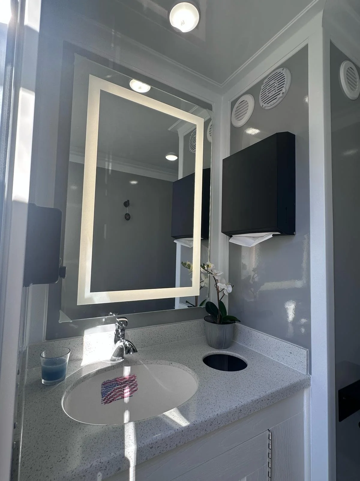 A bathroom vanity with a round white sink, a faucet, a glass with blue liquid, a potted plant, and a backlit mirror. There are wall-mounted black cabinets, a tissue box, and vents on the wall. A ceiling light is also visible.