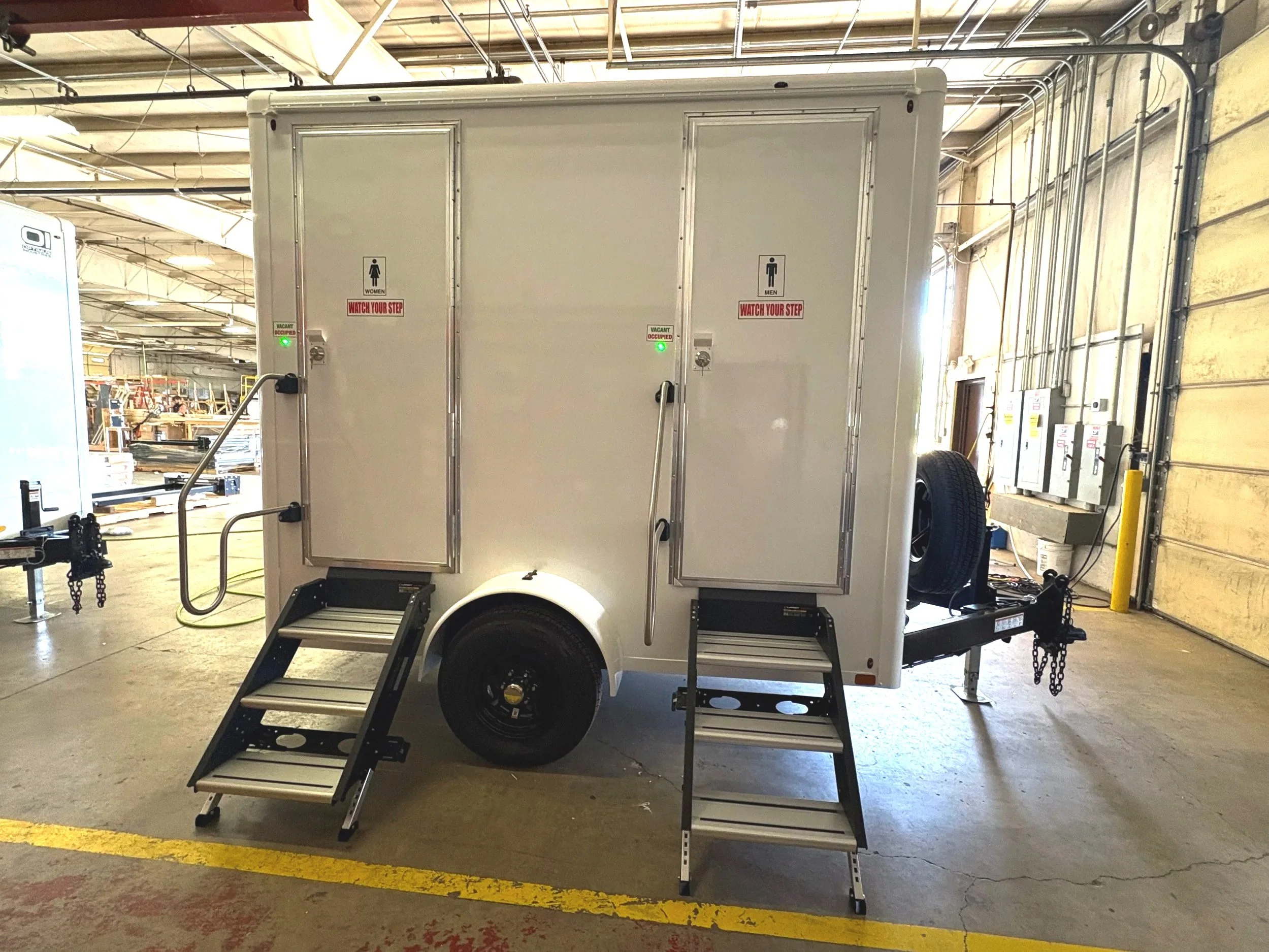White mobile restroom trailer in an industrial warehouse, with steps leading to the entrance, and signs indicating women's and men's restrooms.