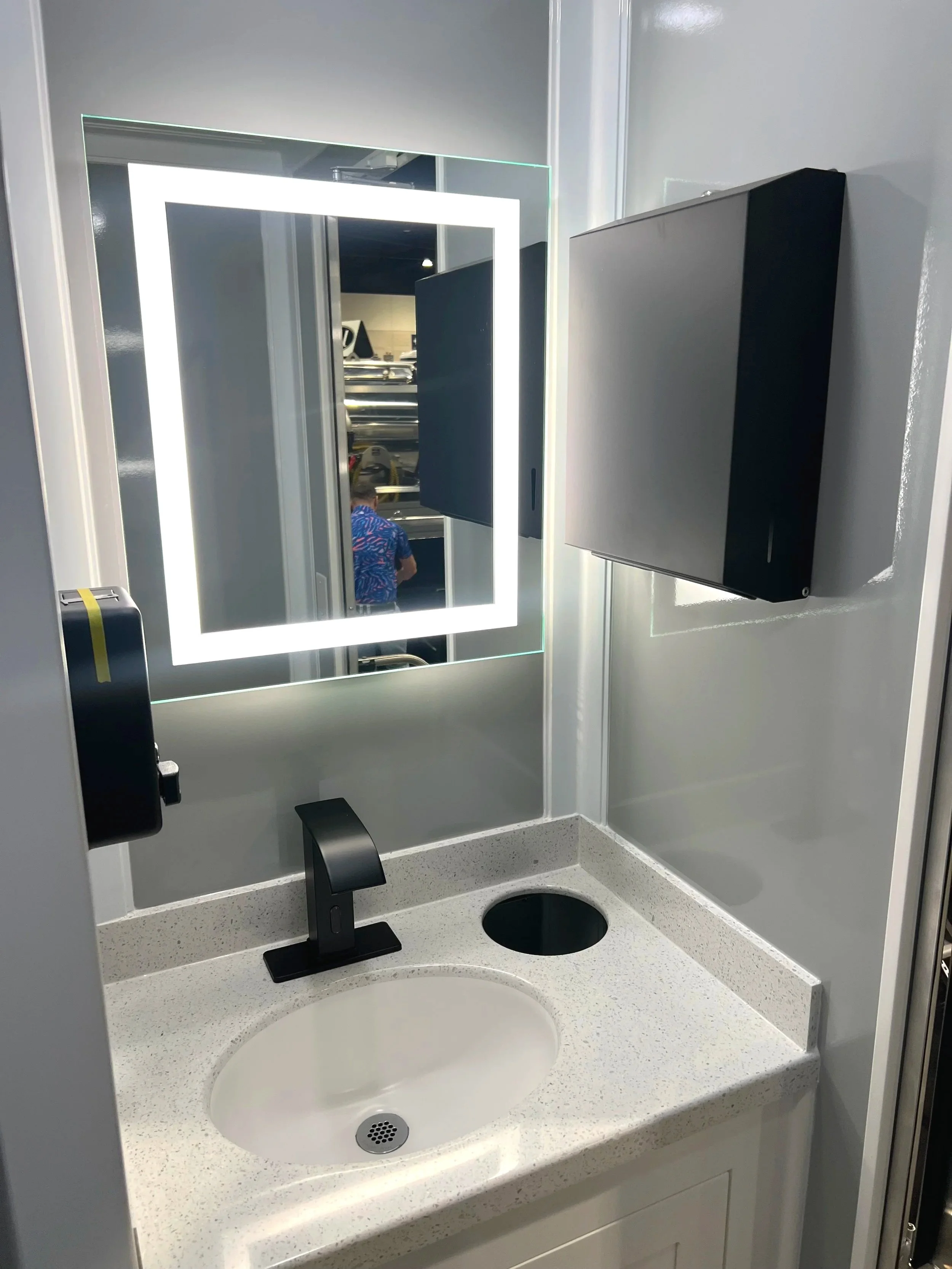 A bathroom sink with a white countertop, a black faucet, and a round sink basin. There is a large mirror with LED lighting, a tissue dispenser on the left, and a black paper towel or hand dryer mounted on the wall. Part of a person wearing a blue patterned shirt is visible in the reflection of the mirror. The wall is painted light gray.