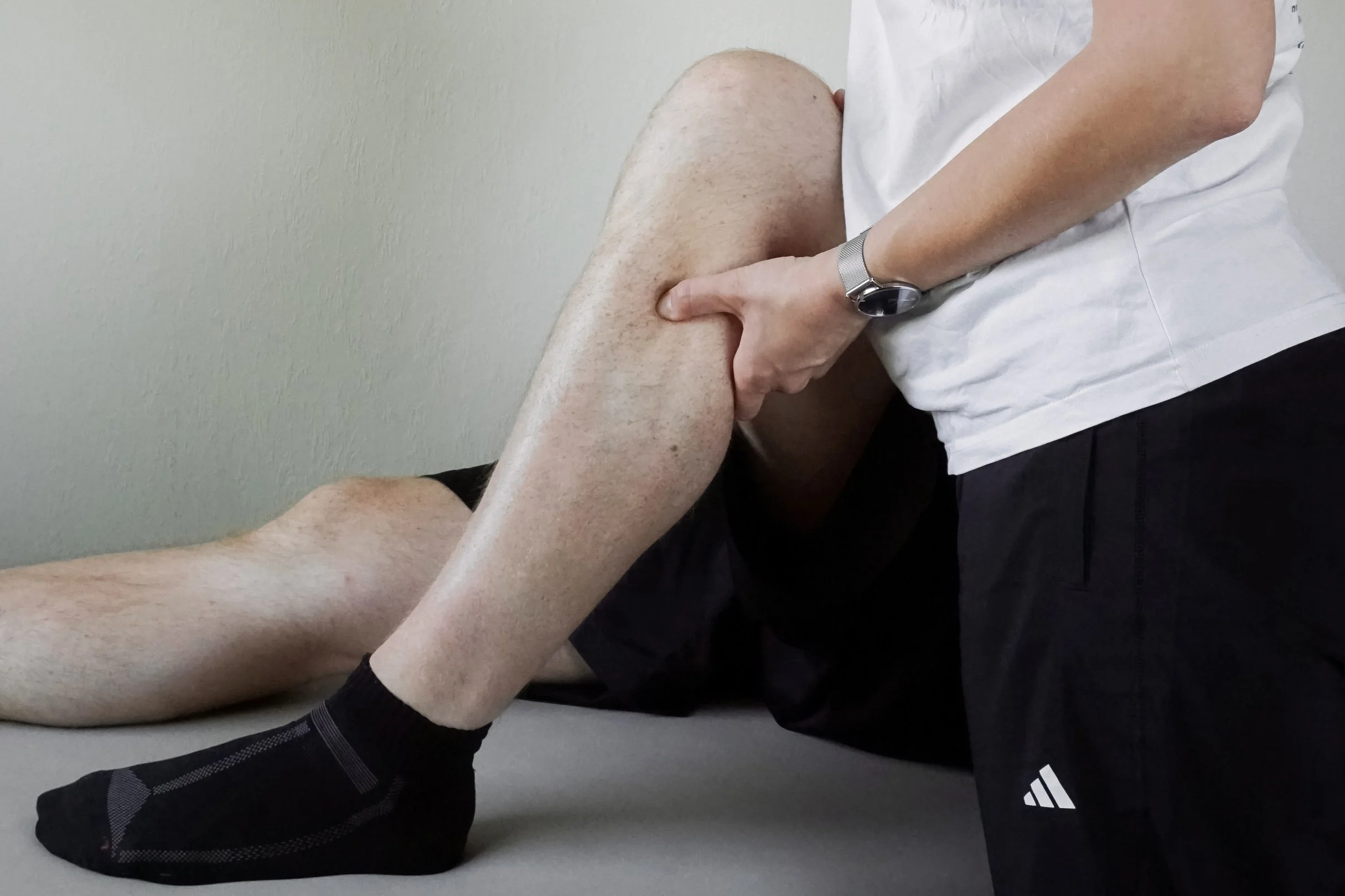 Ein Mensch sitzt auf dem Boden und hält ein ausgestrecktes Bein mit beiden Händen. Es scheint sich um eine Dehnungsübung oder Physiotherapie zu handeln.