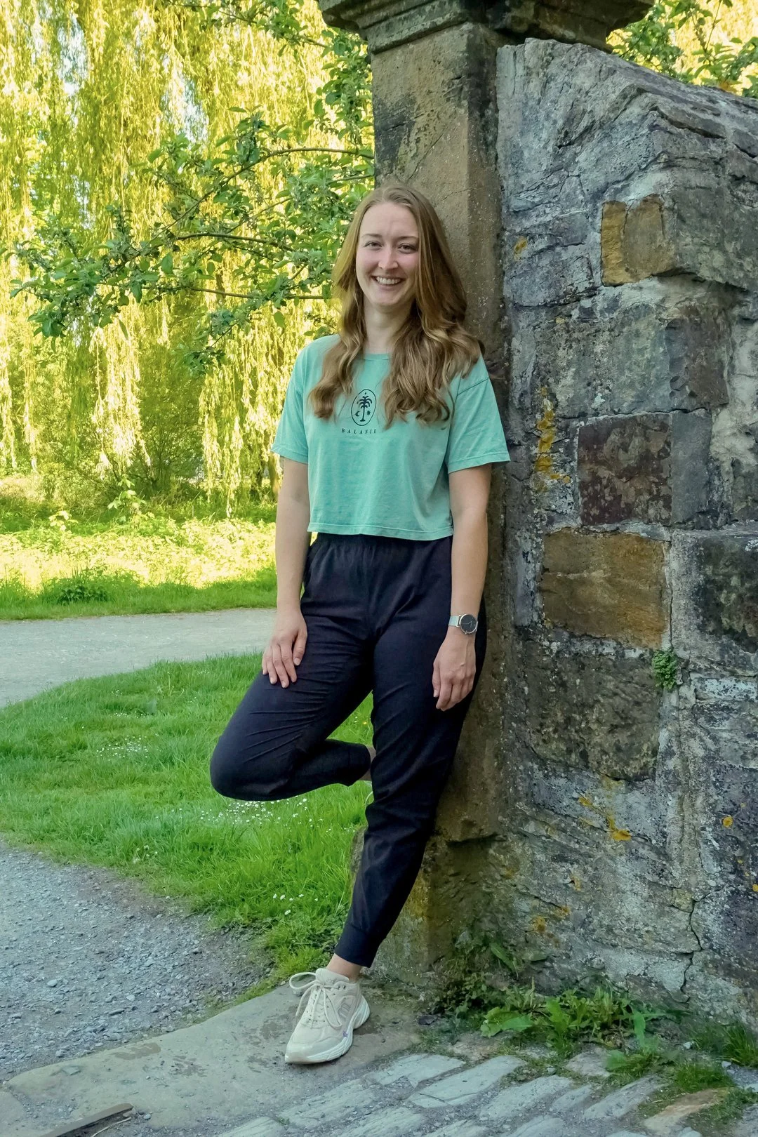 Eine junge Frau mit braunen, langen Haaren, die in einem grünen T-Shirt, schwarzen Hosen und weißen Turnschuhen neben einer alten Steinemauer im Park steht und lächelnd in die Kamera schaut.