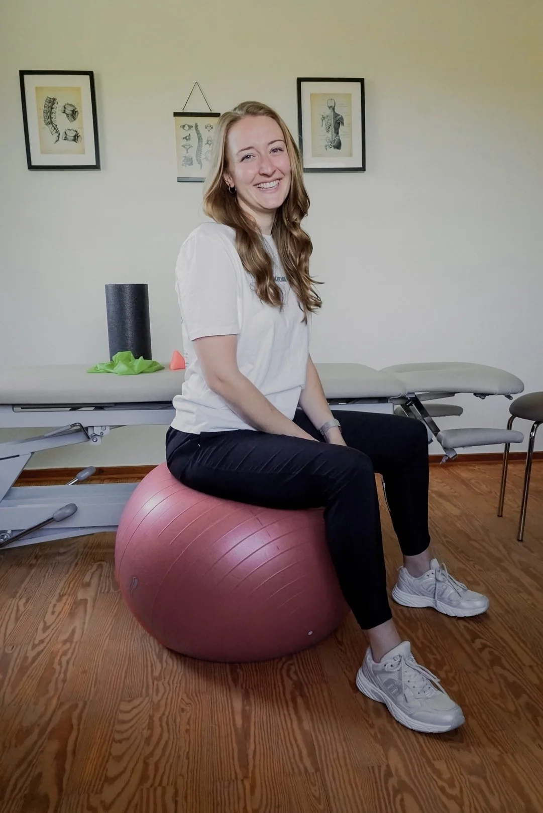 Junge Frau sitzt auf einem roten Gymnastikball in einem Therapie- oder Fitnessraum, im Hintergrund Wandbilder mit Wirbelsäulen- und Muskelzeichnungen.