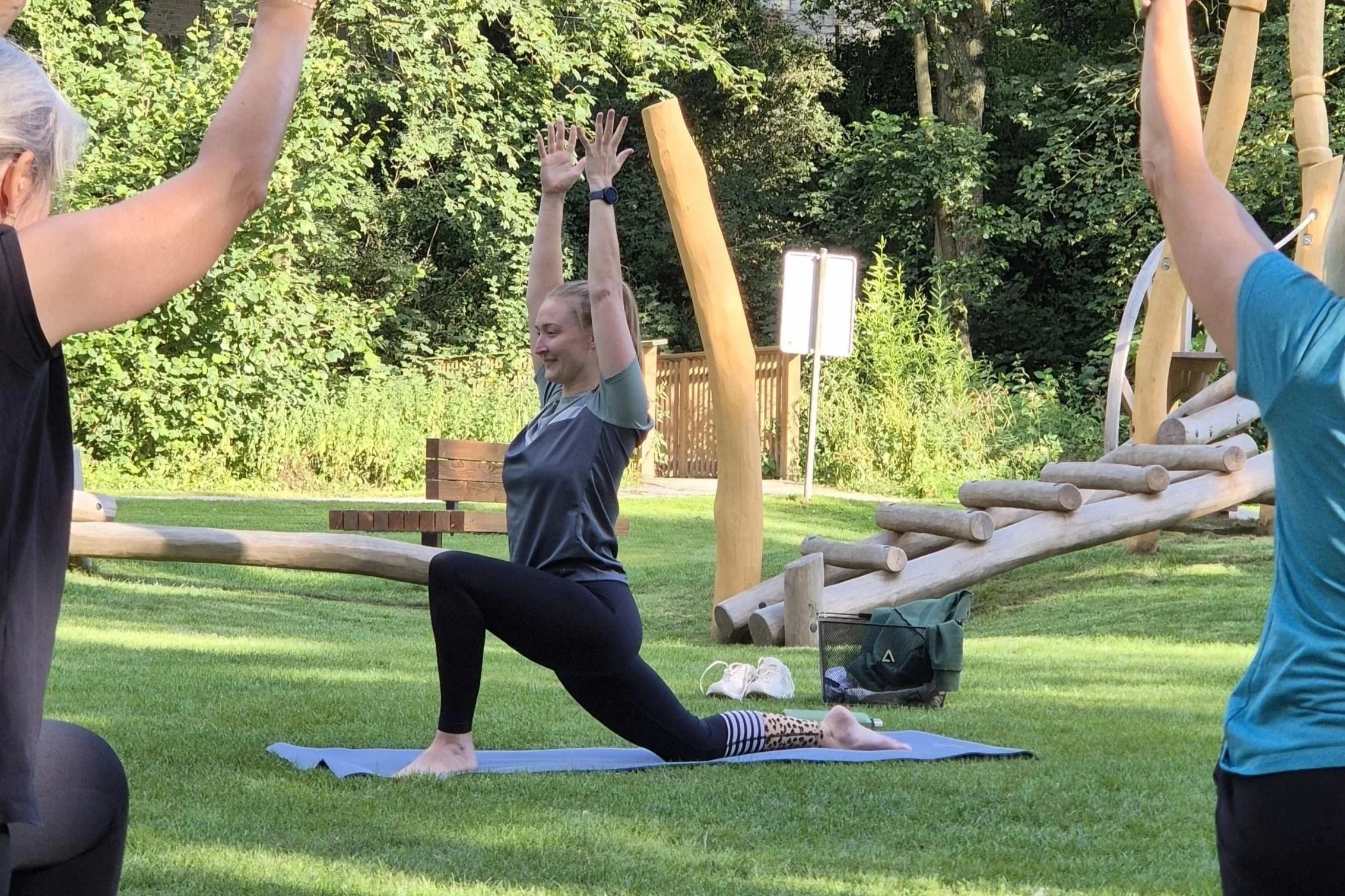 Eine Frau macht Yoga im Freien auf einer Yogamatte, umgeben von Bäumen, während sie in einen Ausfallschritt geht mit nach oben ausgestreckten Armen.