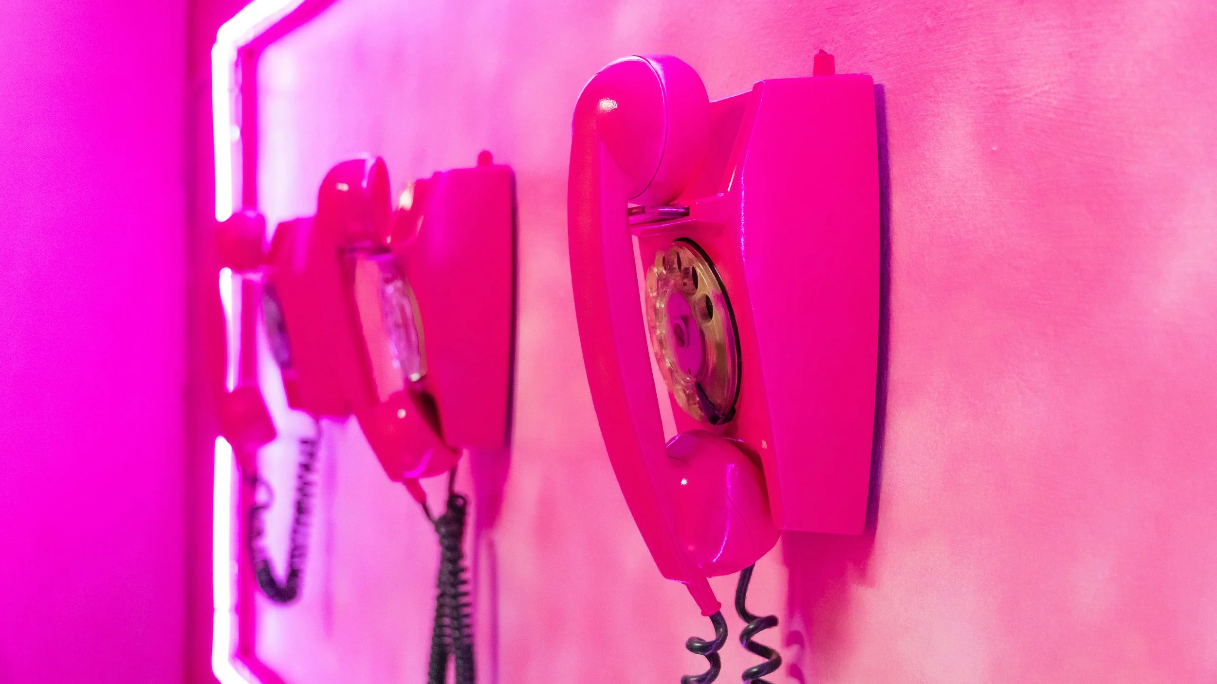 Multiple pink vintage payphones mounted on a pink wall, illuminated by pink neon light.