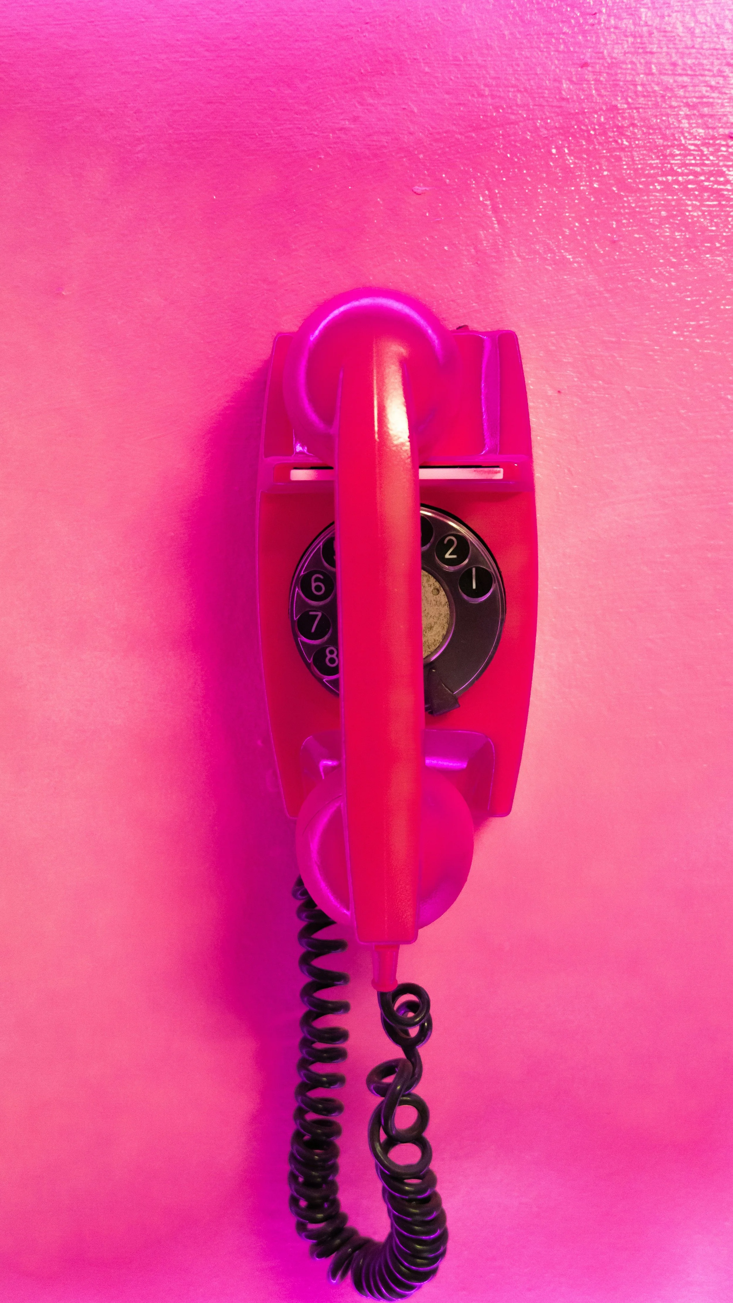 Pink telephone mounted on a pink wall, with a coiled wire and a circular dial with numbered holes.