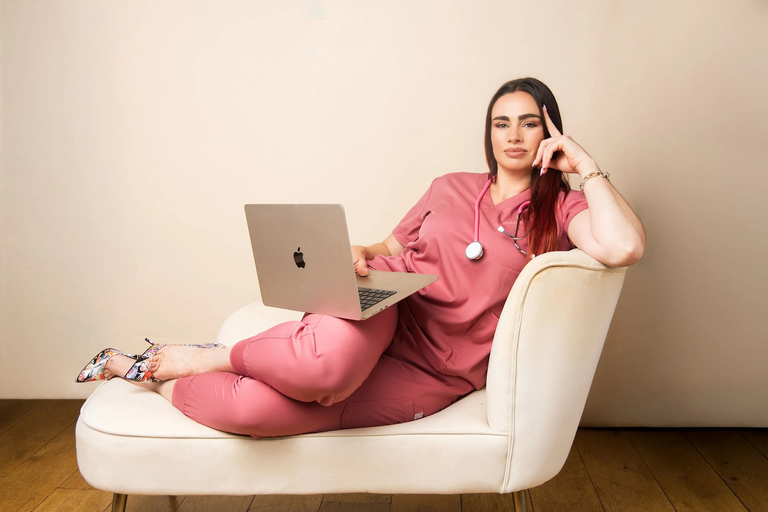 A woman in pink medical scrubs, with a stethoscope around her neck, relaxes on a white sofa while working on a silver laptop. She has long dark hair, and wears floral high heels and a bracelet.