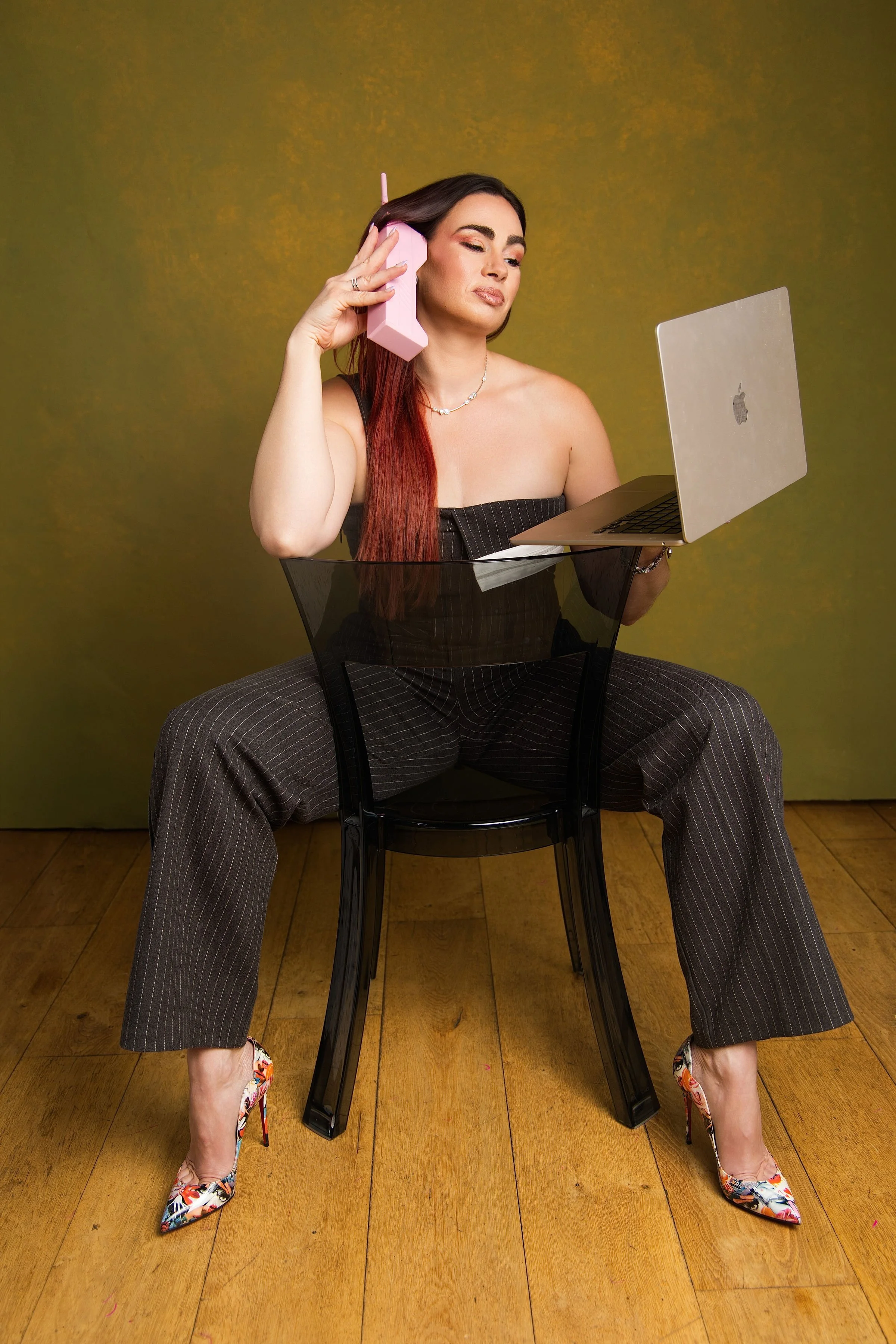 A woman with long dark hair, wearing a black strapless top, pinstriped pants, floral high heels, and a necklace, seated on the floor with her legs apart against a green background, holds a pink rotary phone to her ear with a focused expression, while holding a silver laptop on her lap.