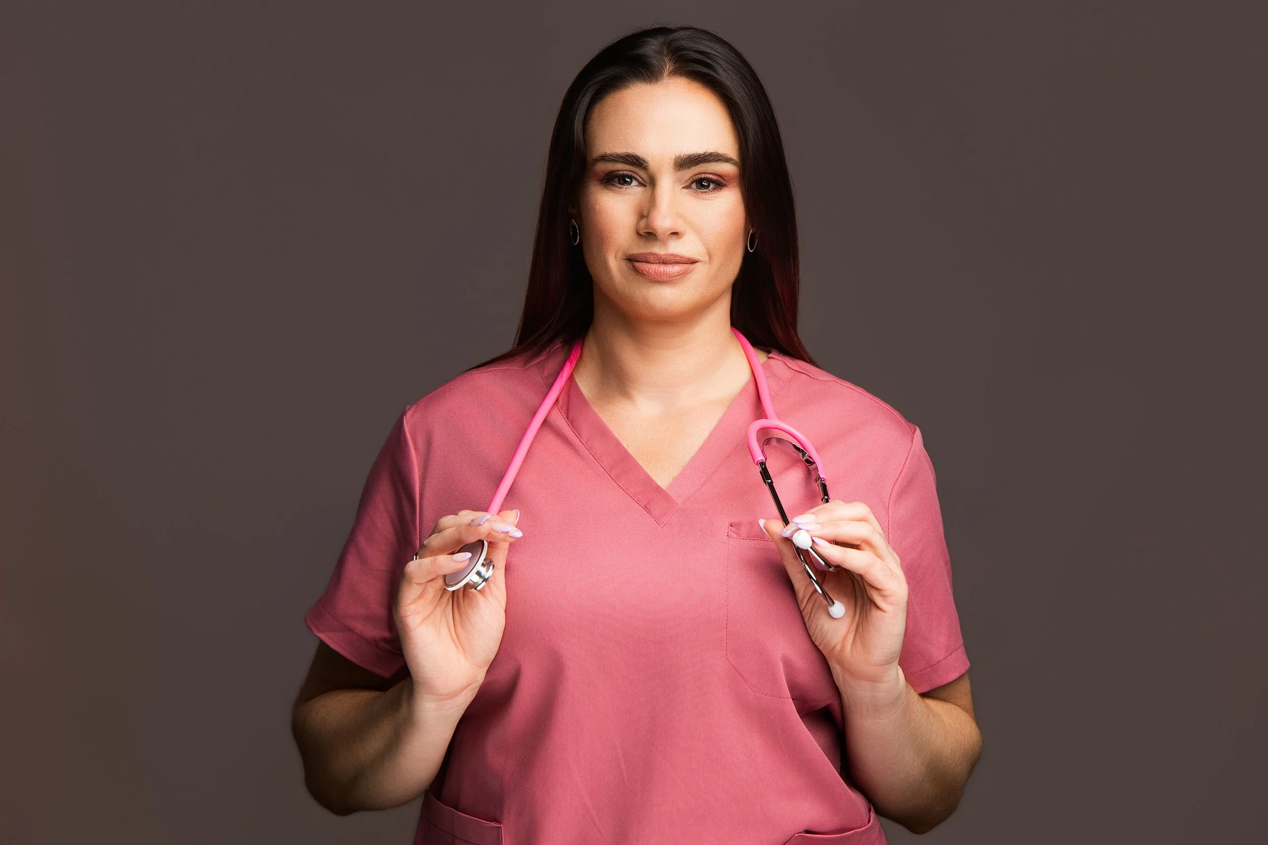 A female healthcare professional with dark hair, wearing pink scrubs and holding a pink stethoscope around her neck, standing against a dark gray background.