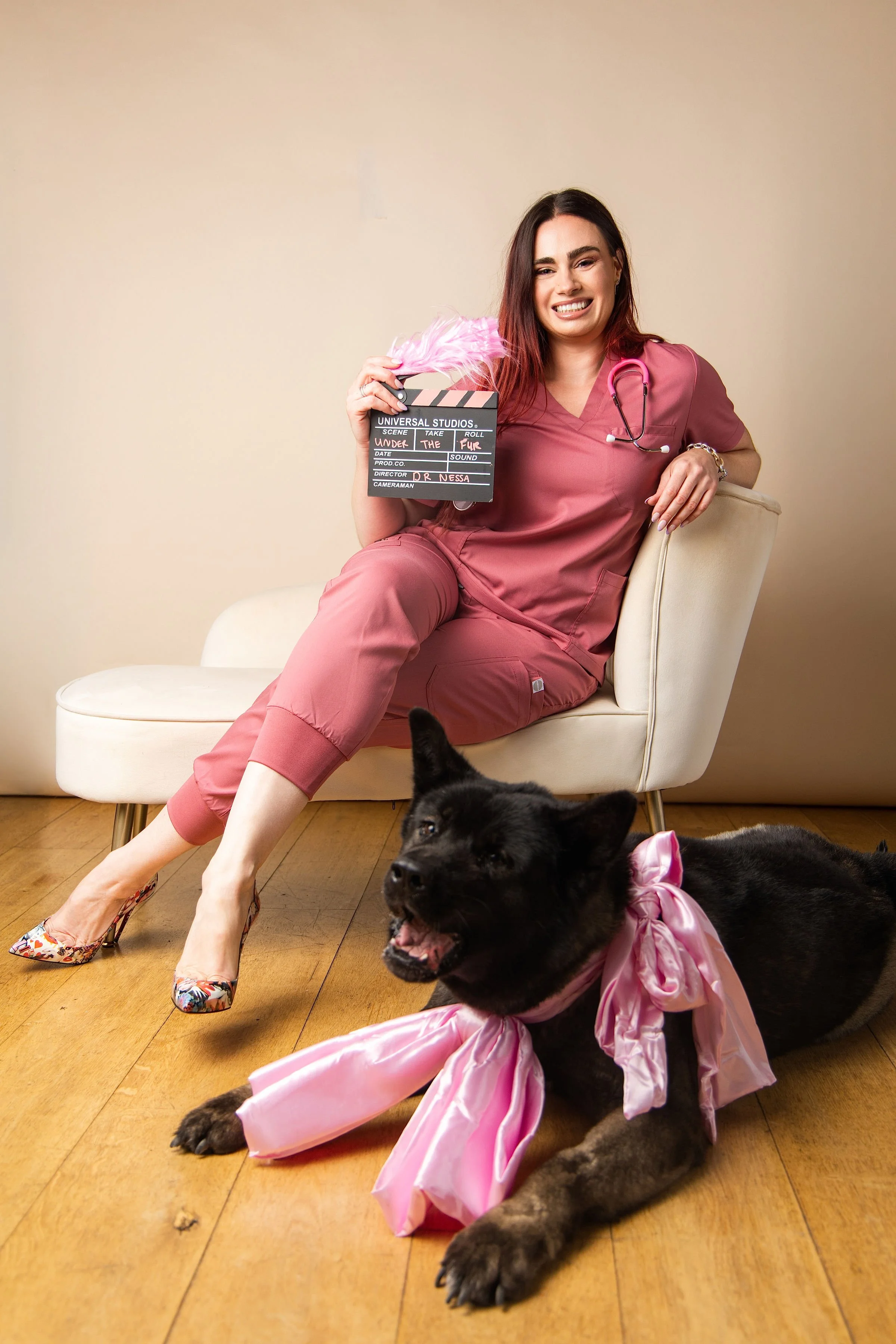 A woman in pink scrubs sitting on a white chair, holding a film clapboard and smiling. She has a stethoscope around her neck and is wearing floral high heels. A black puppy with a pink bow on its neck is lying on the wooden floor in front of her, panting with tongue out.