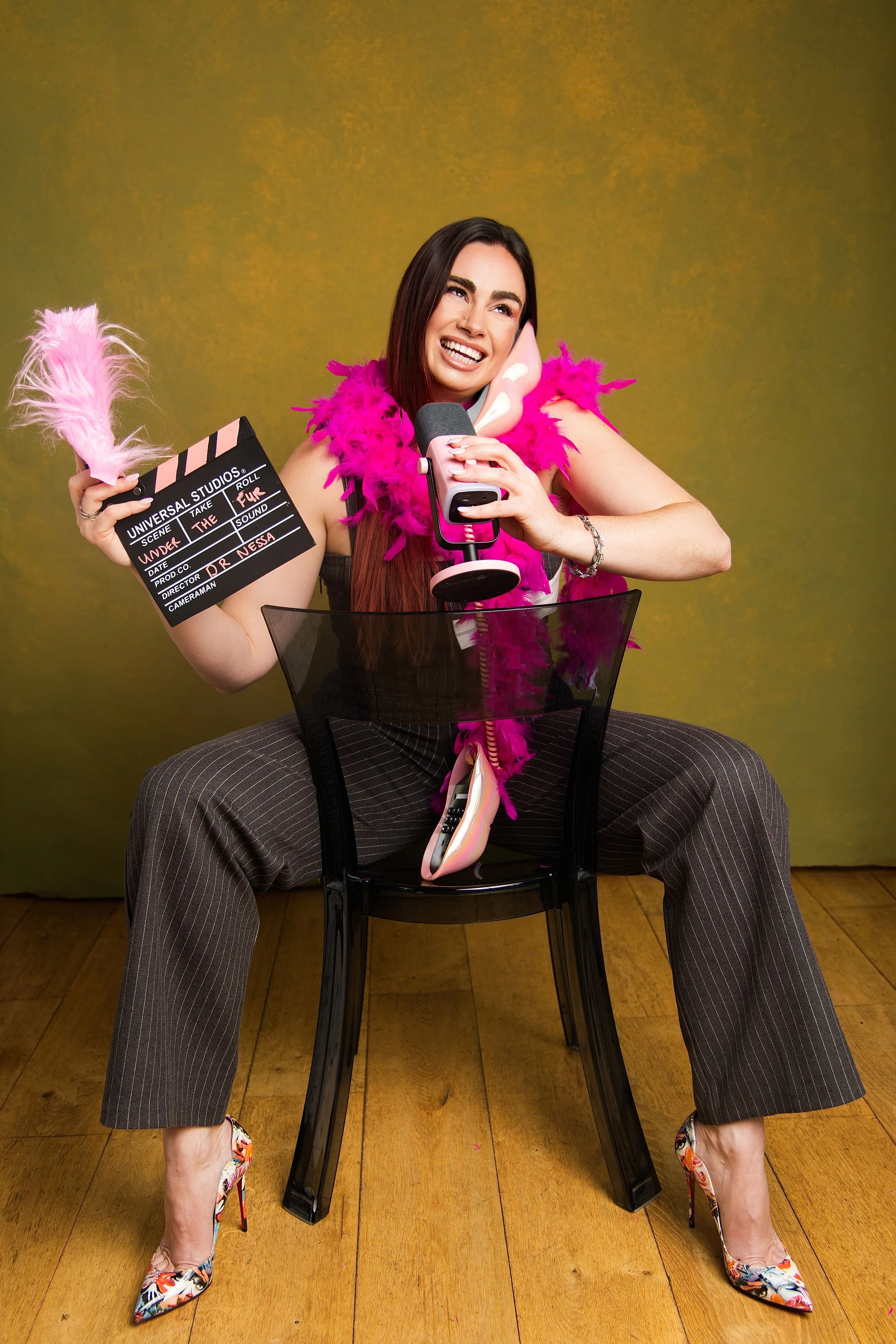 Woman with long dark hair, wearing a pink feather boa, striped pants, colorful high heels, sitting on a black chair with legs apart, holding a clapperboard in one hand and a pink microphone with a pig face in her other hand, smiling and talking into the microphone, against a yellow background.