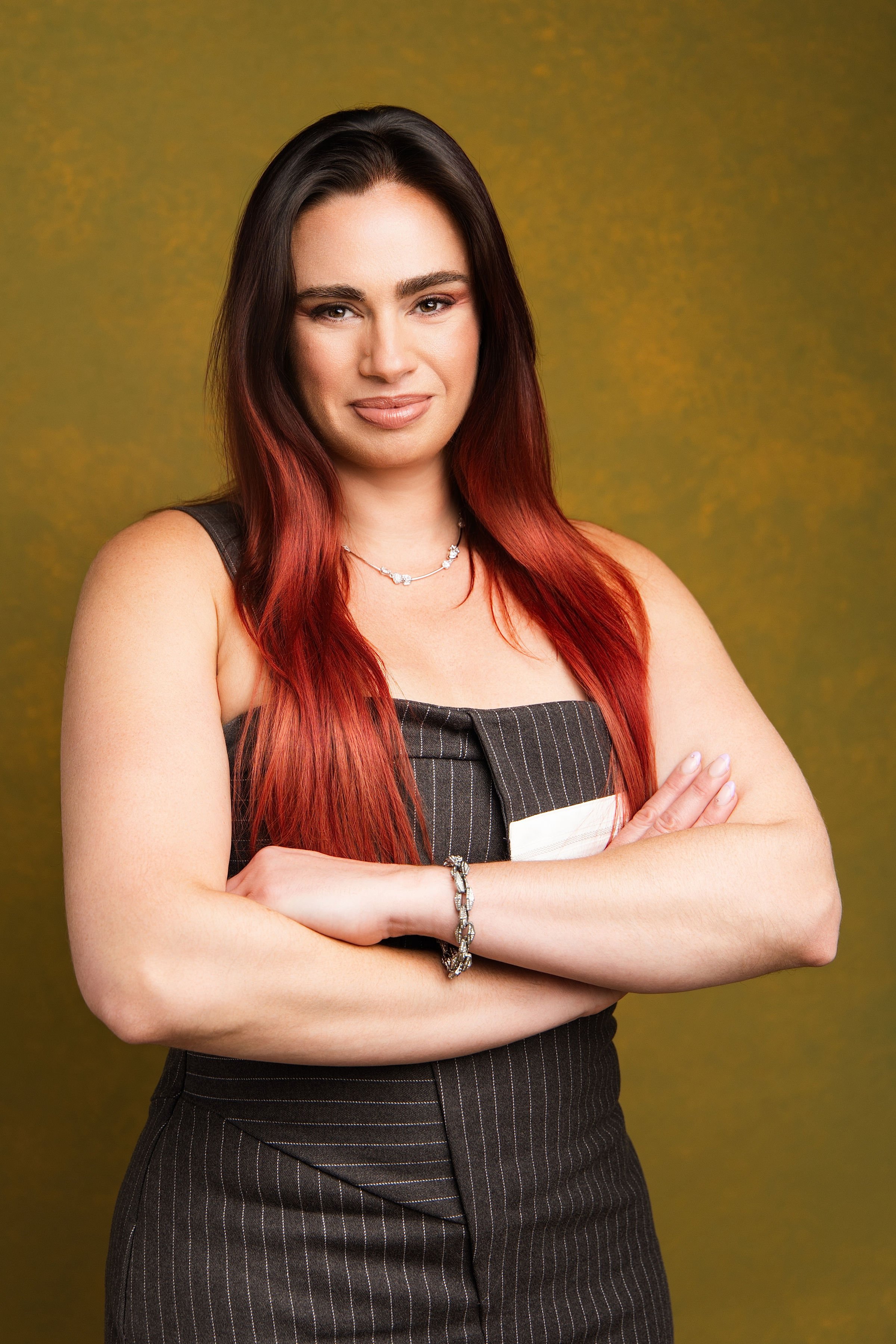 A woman with long, dark red hair, wearing a black sleeveless top and pinstriped pants, standing with her arms crossed against a yellow background.