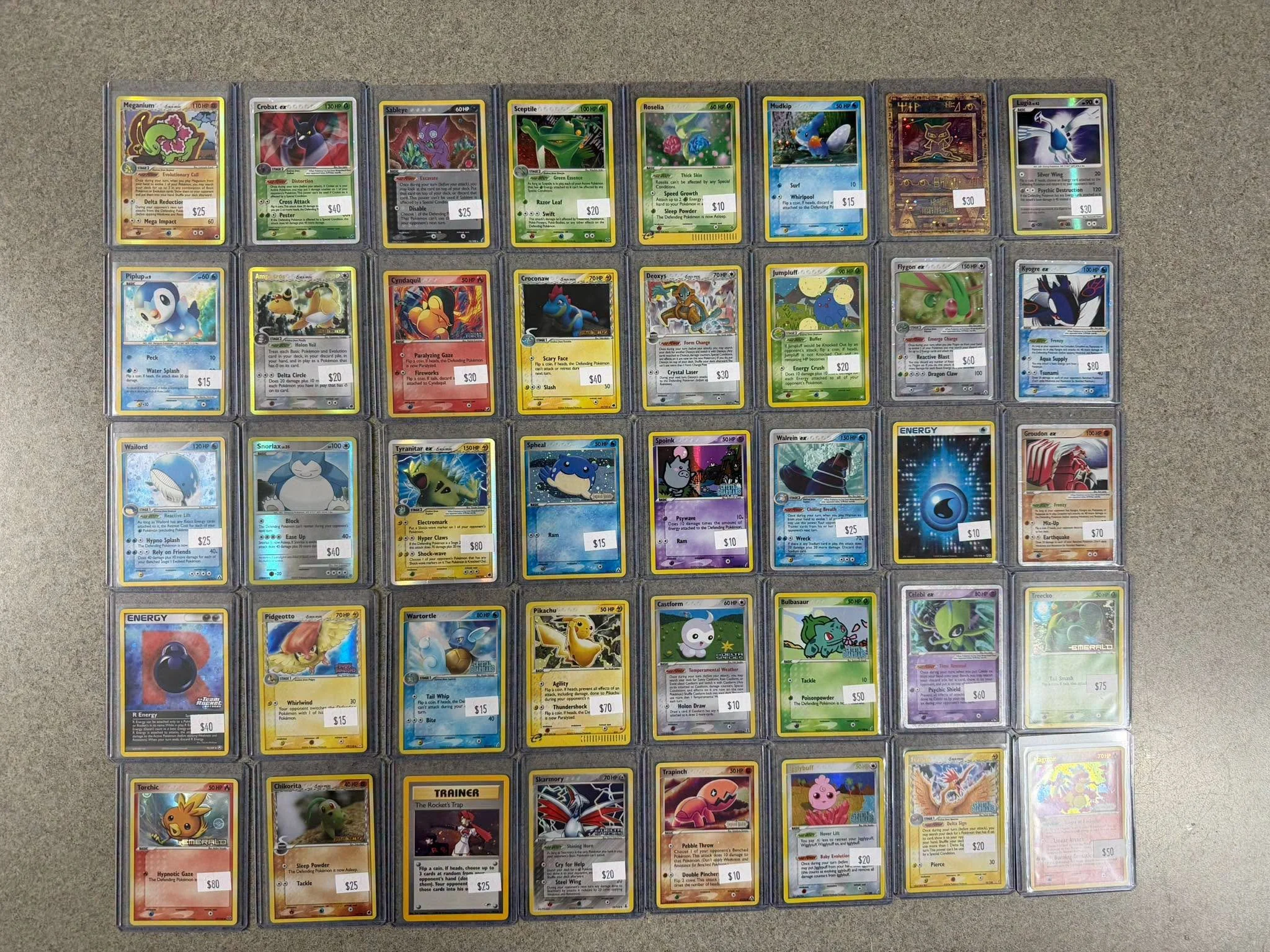A collection of Pokémon trading cards arranged in a 4 x 7 grid on a gray surface. The cards vary in design, featuring different Pokémon characters and some with prices and labels.