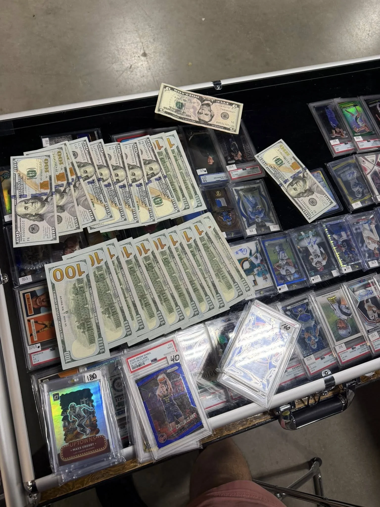 A display case with multiple stacks of US $100 bills, some loose bills, and various collectible cards inside protective cases, with a gray floor background. Buying a sports card collection at a card show