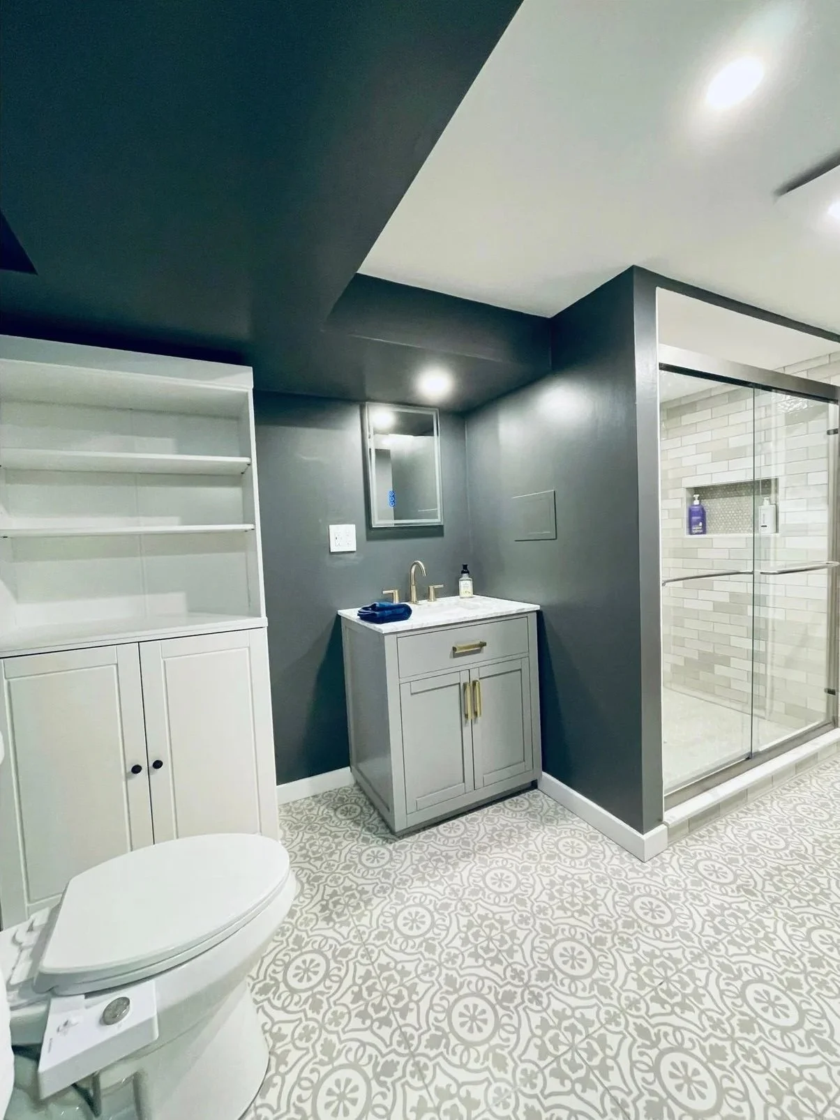 A modern bathroom featuring a white toilet, a white and gray vanity with a marble top and gold hardware, a gray wall with a mirror and a soap dispenser, a white storage cabinet, and a glass-enclosed walk-in shower with beige subway tiles and purple and white bath products inside.