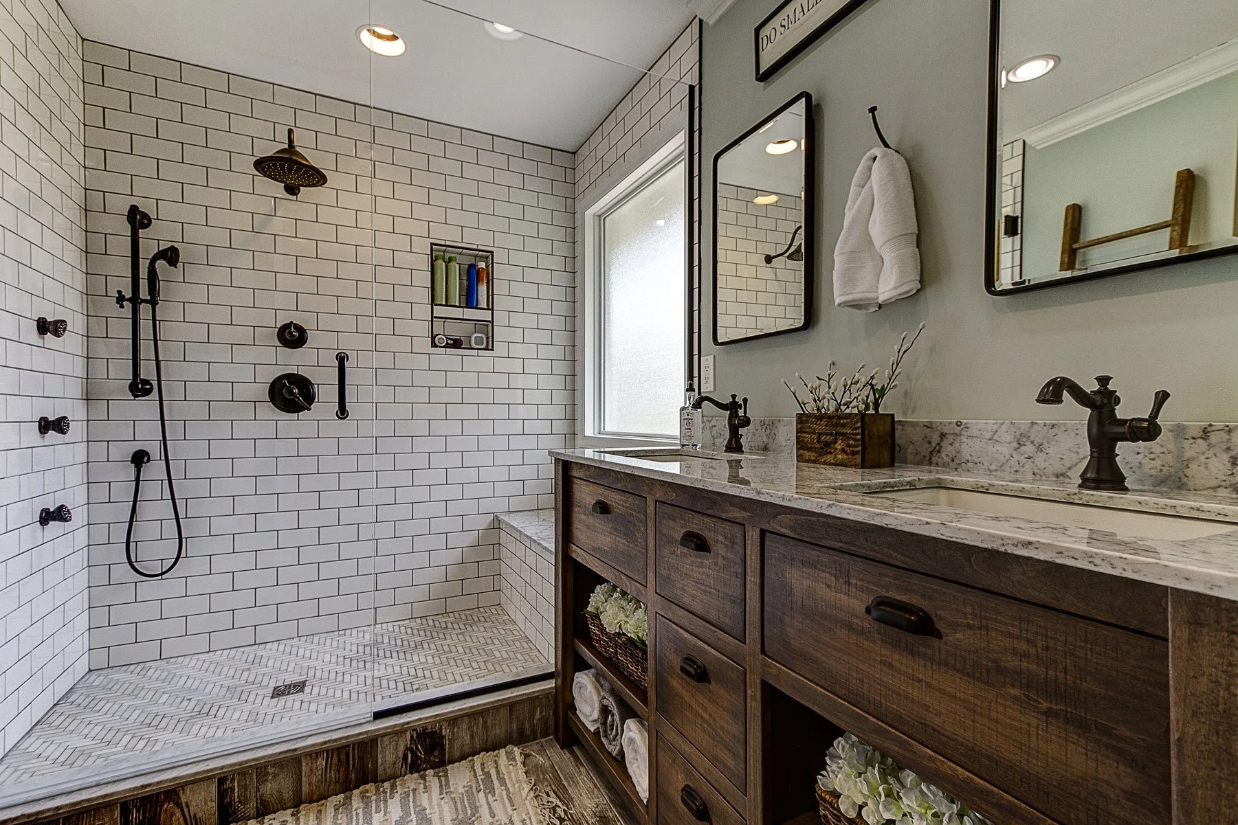 Modern bathroom with walk-in shower featuring white subway tiles, black fixtures, and niche for toiletries. Wooden vanity with granite countertop, undermount sink, and mirrors. Decor includes a small flower arrangement and towels.