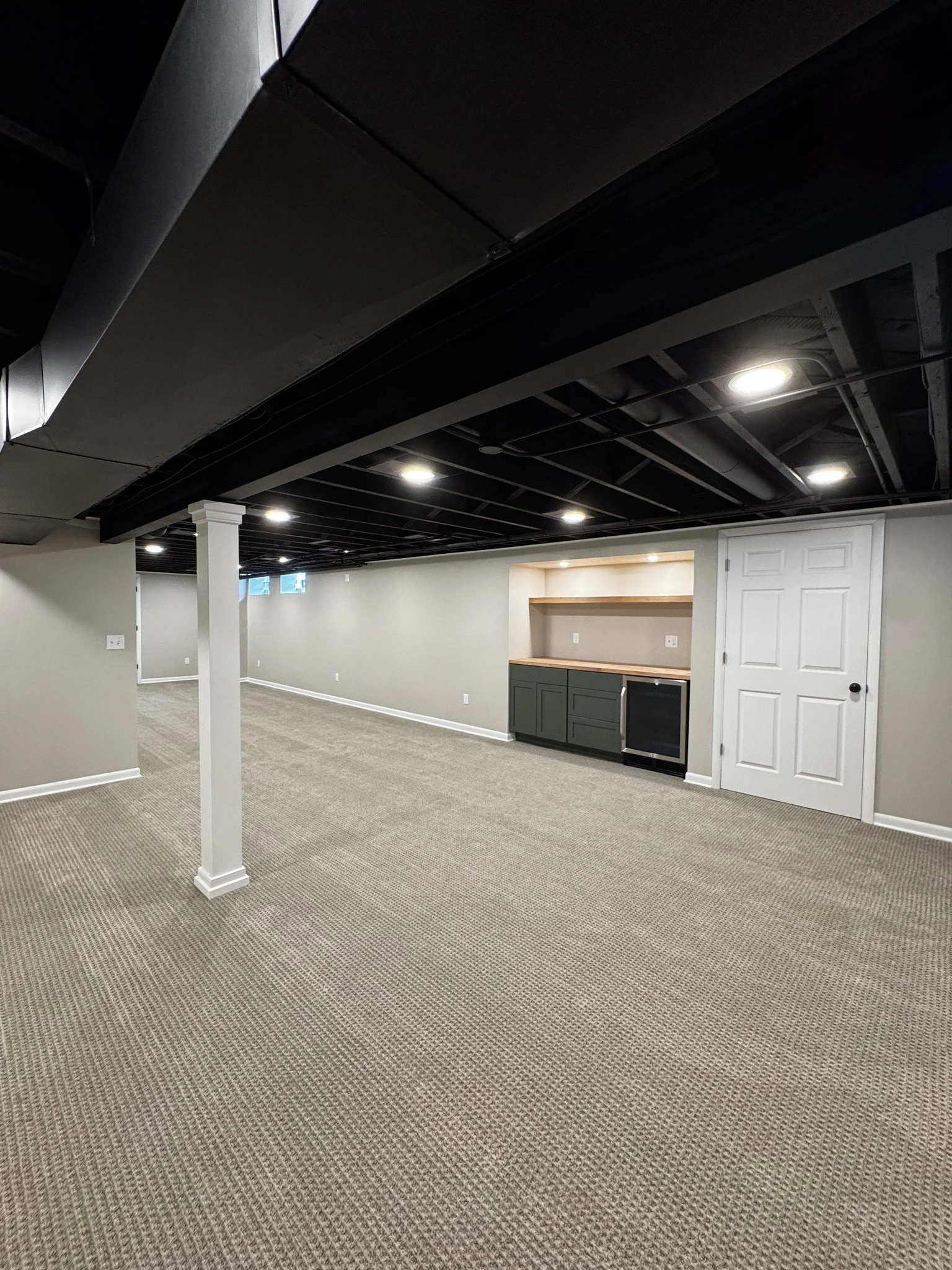 Unfinished basement with beige carpet, white walls, black ceiling beams, and a small built-in cabinet or bar area with a mini fridge and open shelving.