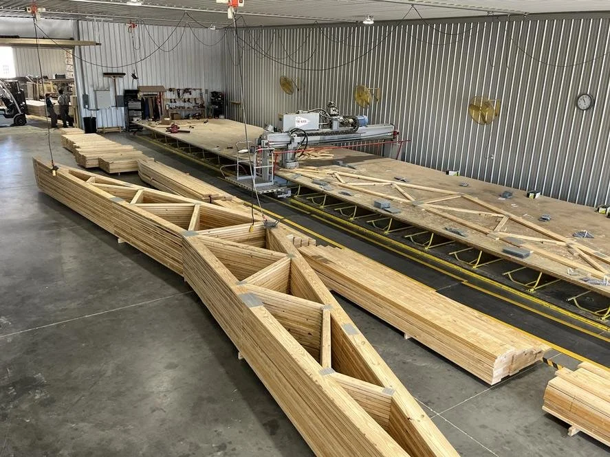Indoor woodworking shop with large wooden boat or structure in progress, surrounded by stacks of lumber, tools, and machinery.