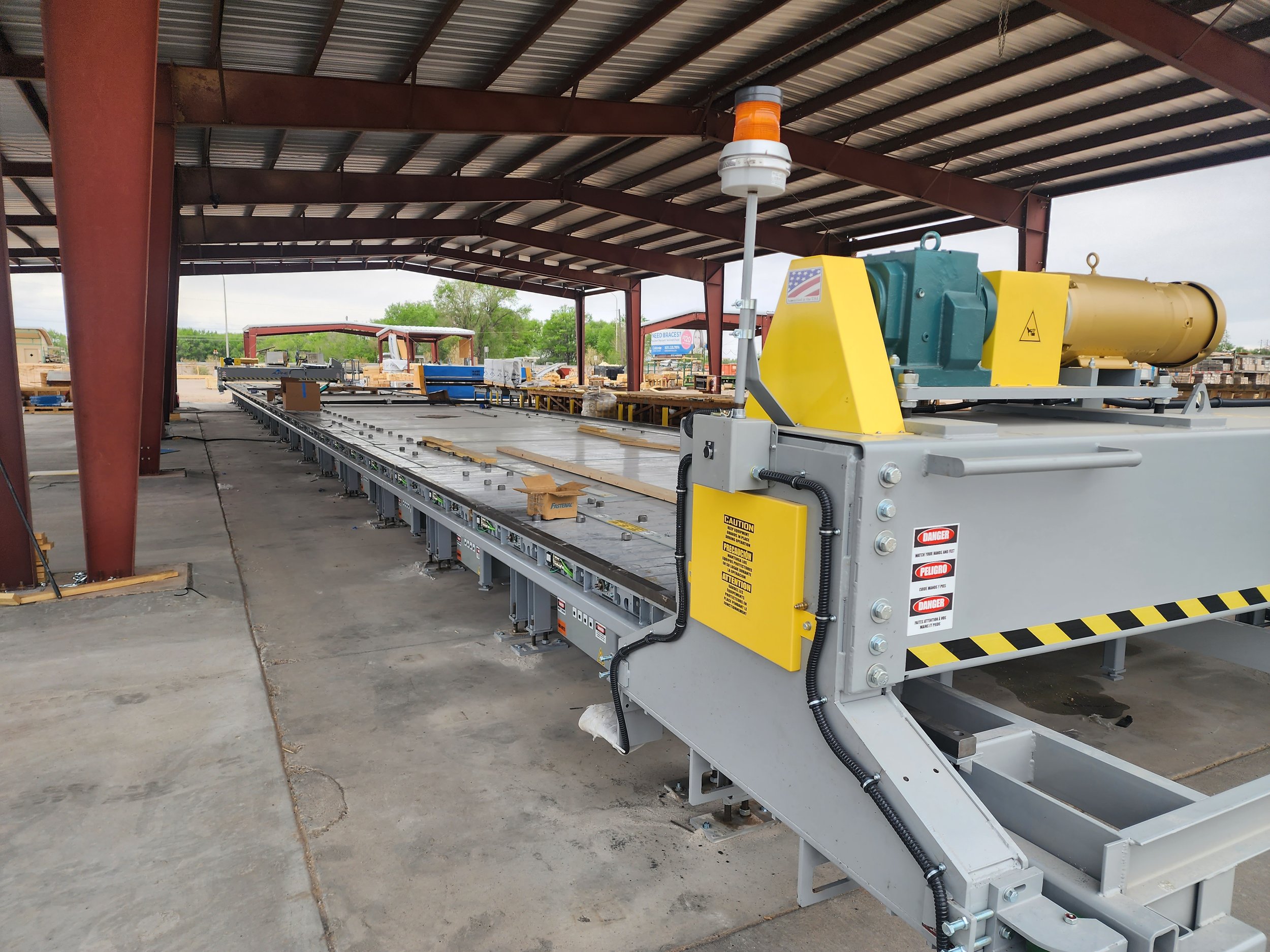 Industrial manufacturing or processing equipment situated outdoors under a metal-roofed shelter with trees and fencing in the background.