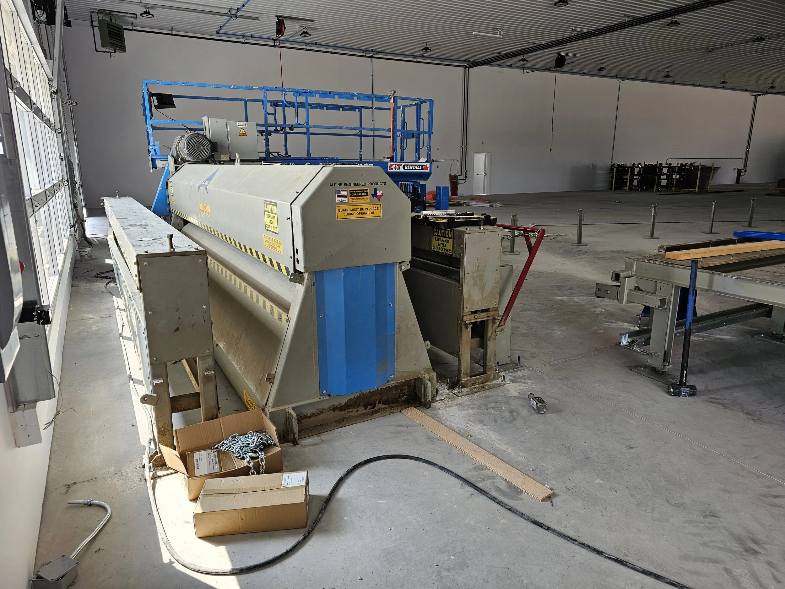 Construction or industrial equipment inside a warehouse or garage, including a large blue scissor lift platform, a machine with caution and warning labels, a cardboard box with chains, and various tools and materials scattered on the floor.