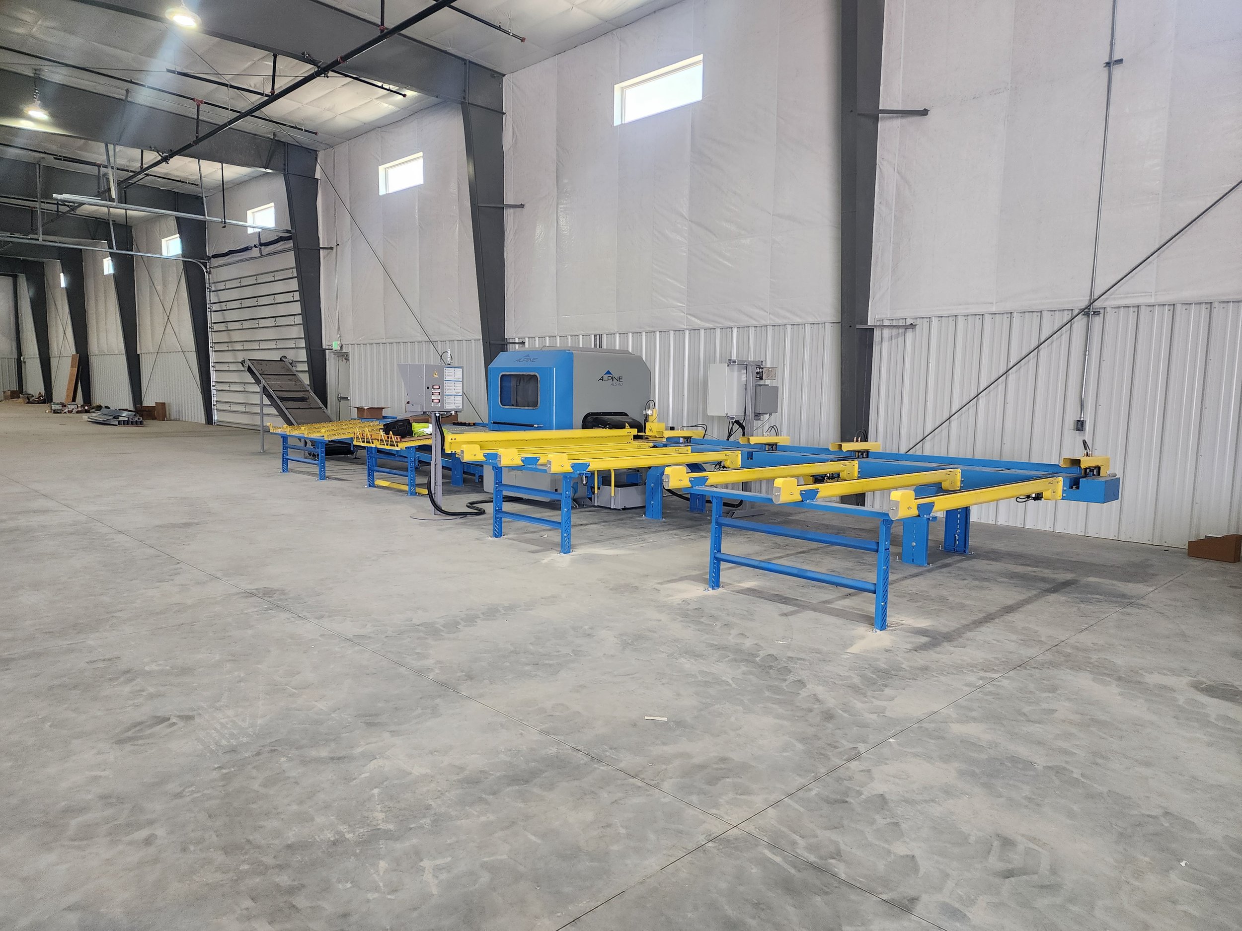 Interior of a large industrial warehouse with metal walls and ceiling, featuring small rectangular windows high on the walls. There are blue and yellow conveyor or storage racks in the foreground, and some equipment and miscellaneous items along the back wall.