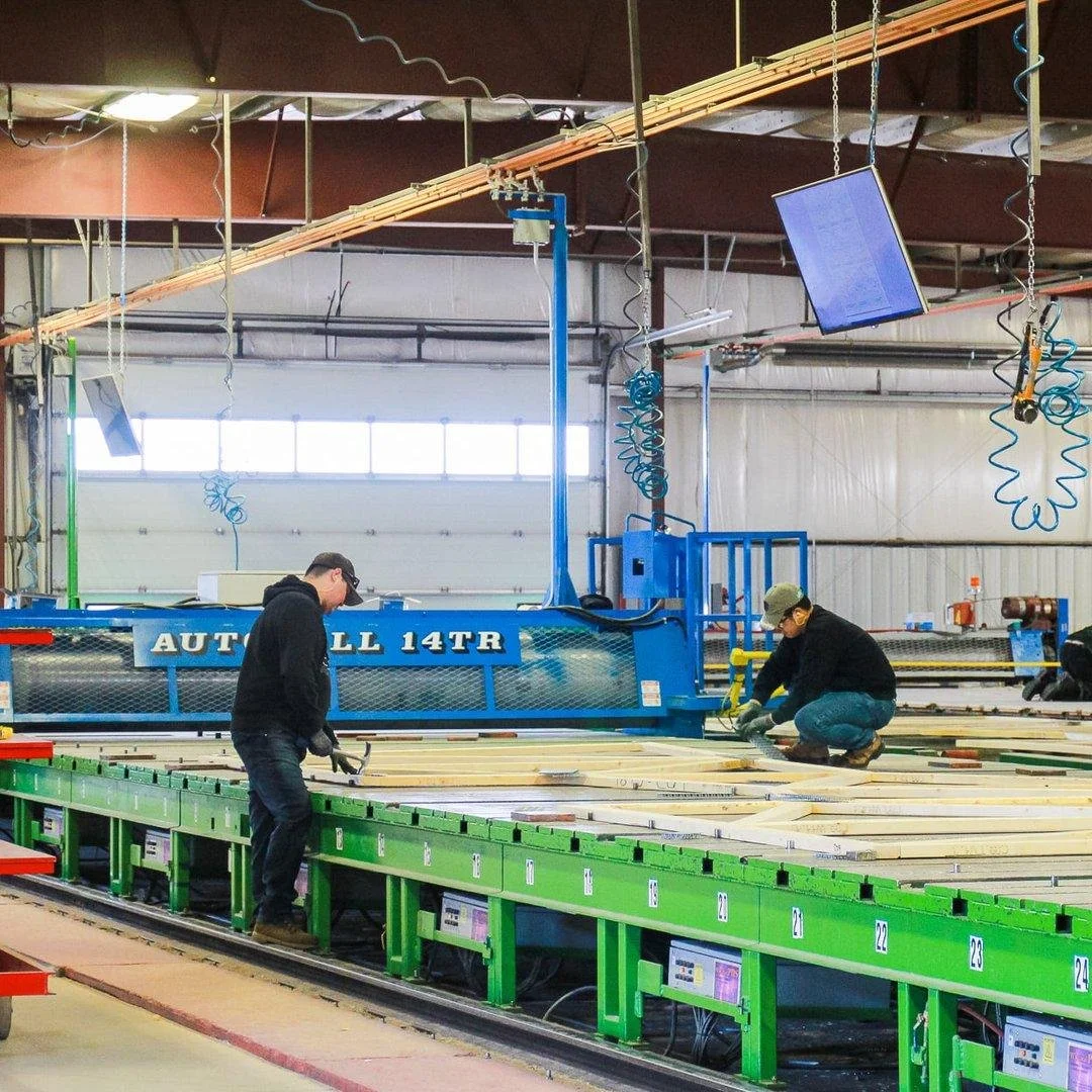 Two workers in a large industrial workshop are laying out wood pieces on a green and metal surface for construction. The workshop has various hanging equipment and screens suspended from the ceiling.