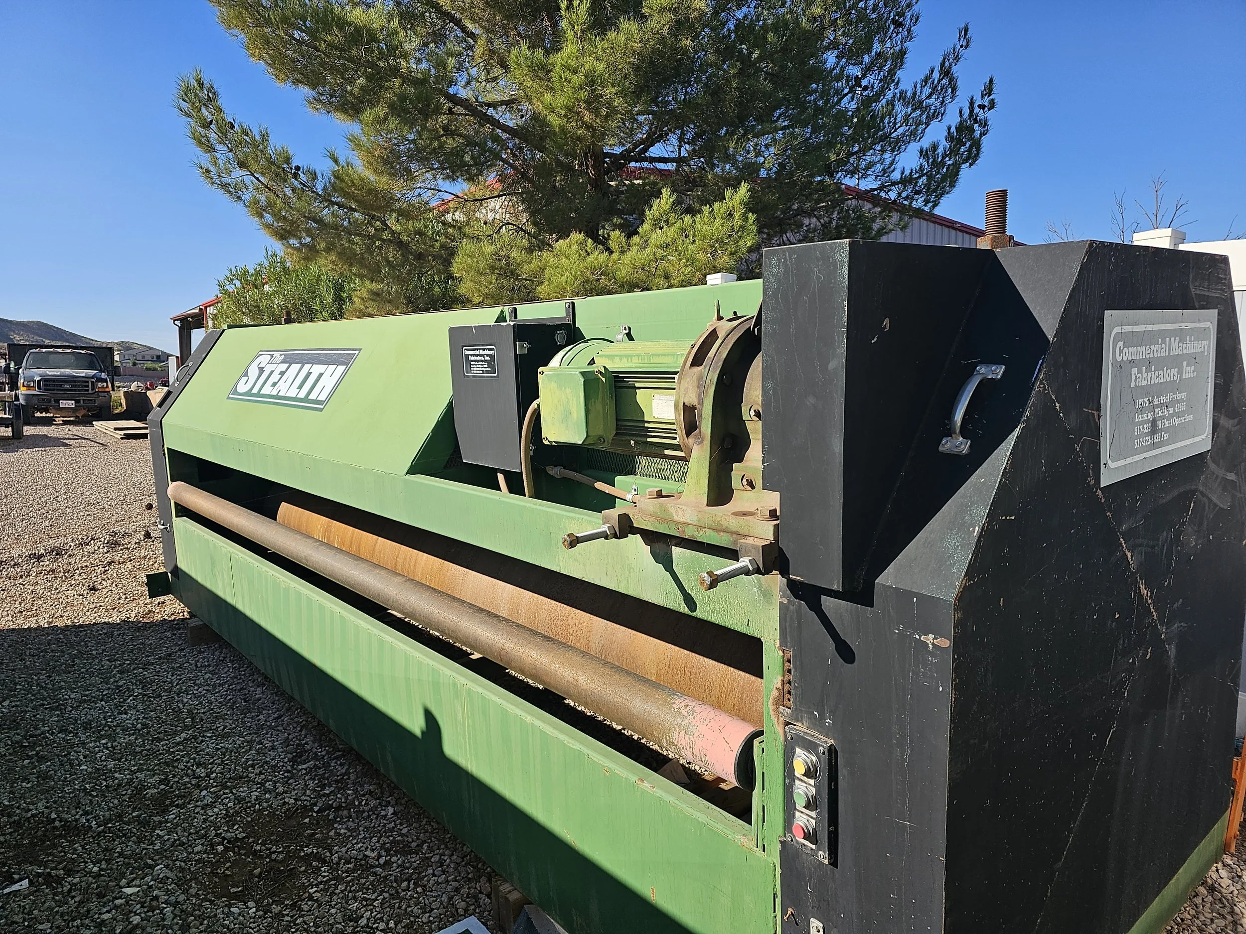 A large green industrial machine with the brand name 'The Stealth' painted on the side, outdoors on a gravel surface with a background of trees, a building with a red roof, and a clear blue sky.