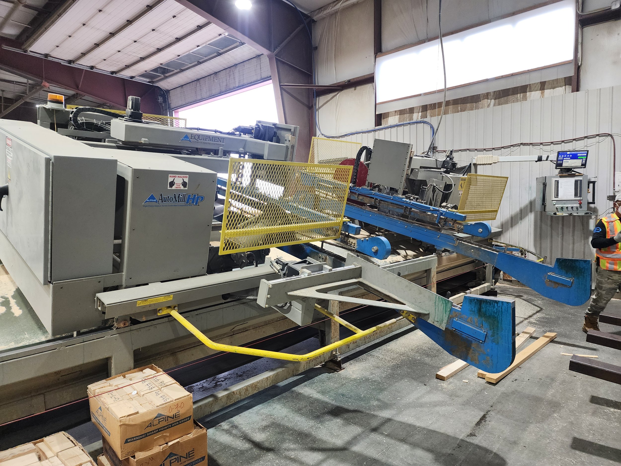 Industrial manufacturing equipment in a warehouse, including conveyor systems, control panels, and safety barriers, with a worker visible in safety gear at the bottom right.