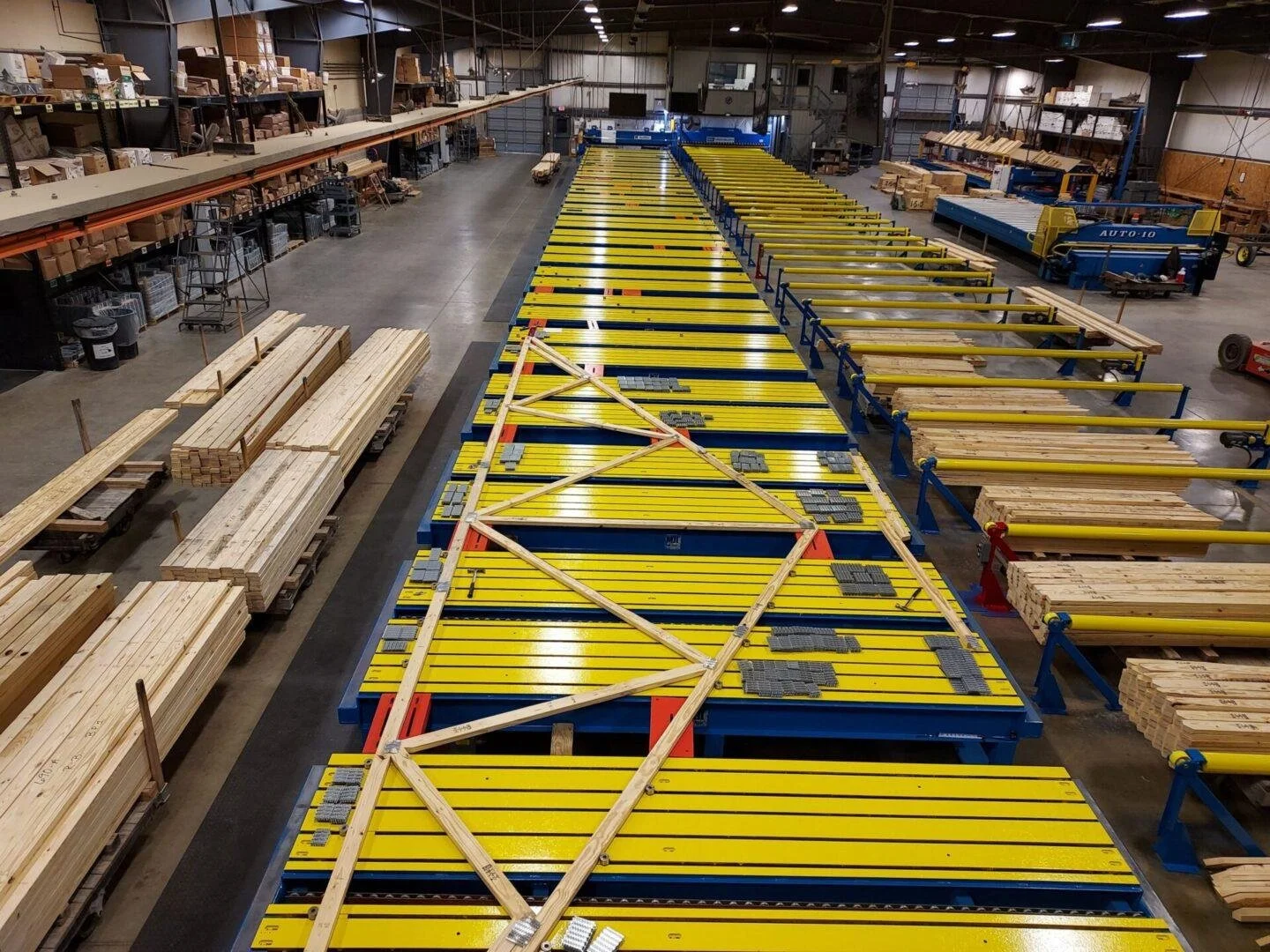 Industrial warehouse with stacks of lumber, large yellow machinery, and wood-cutting equipment.