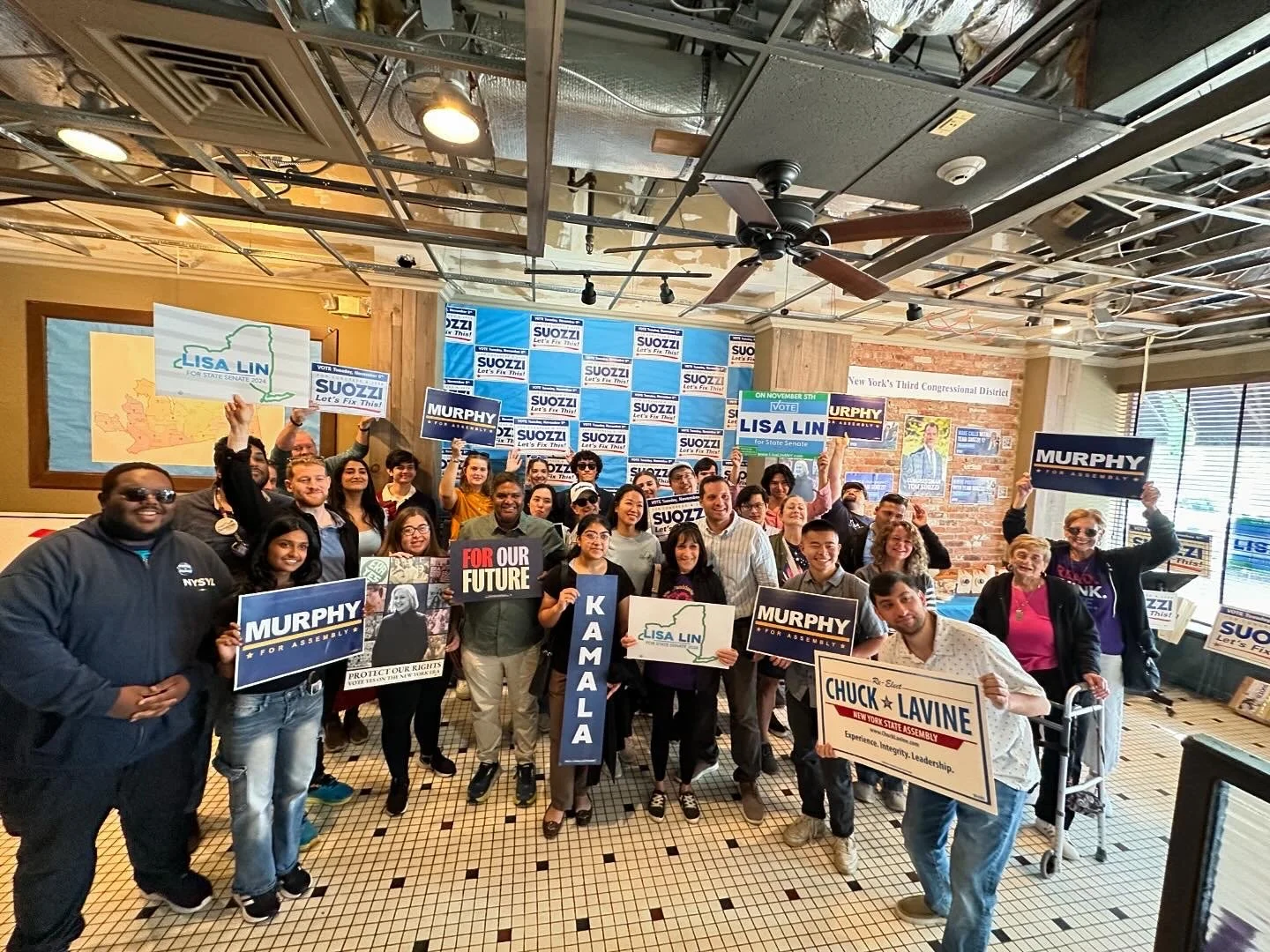 A diverse group of people holding campaign signs for Lisa Lin, Murphy, Kamala, and Chuck LaVine, gathered indoors for a political event with campaign posters on the wall, including a map of New York.