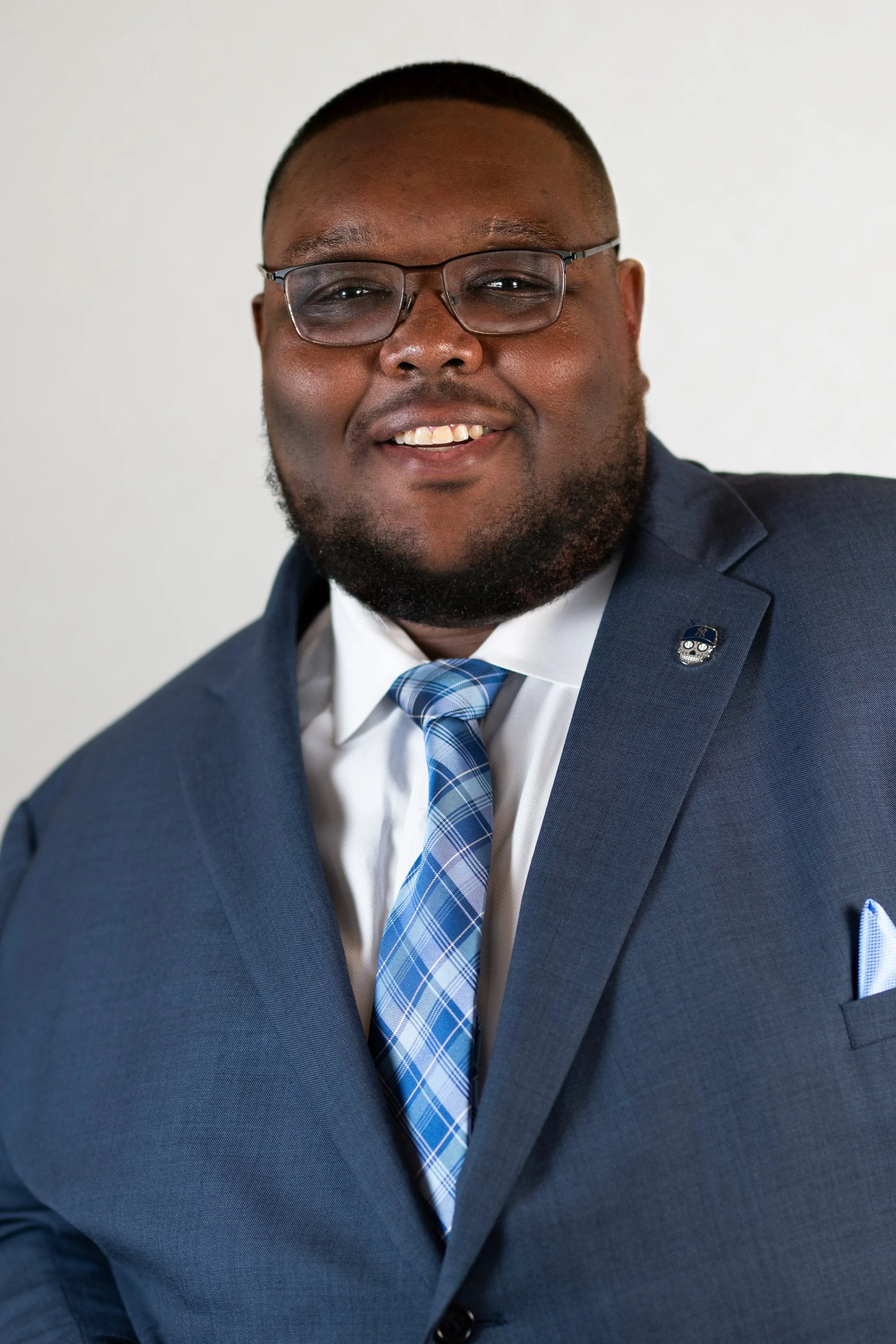 A smiling Black man in a navy blue suit, white shirt, and blue plaid tie, wearing glasses, with a badge on his lapel, against a plain white background.