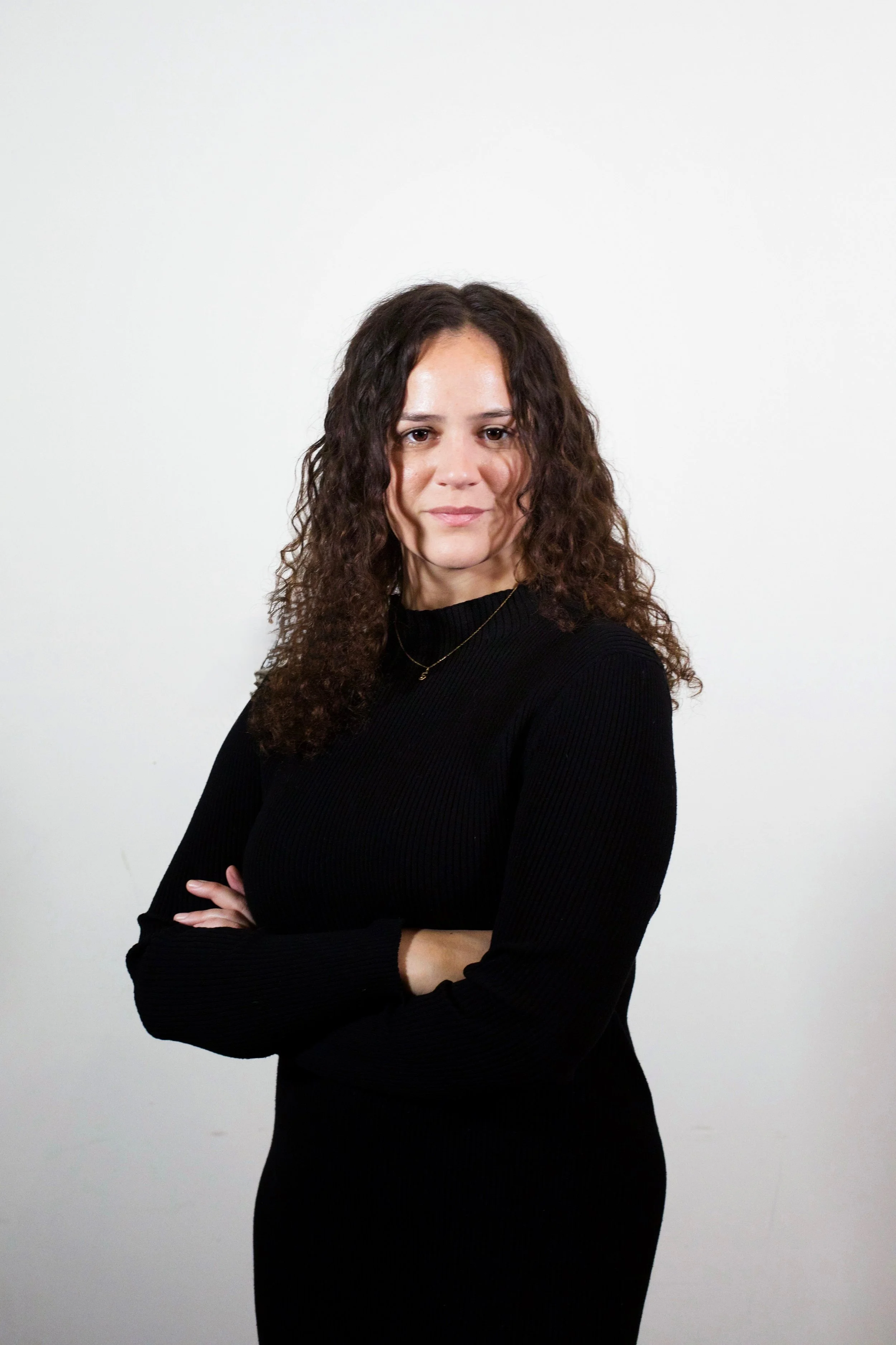 A woman with curly brown hair standing with her arms crossed against a plain white background, wearing a black long-sleeve top.